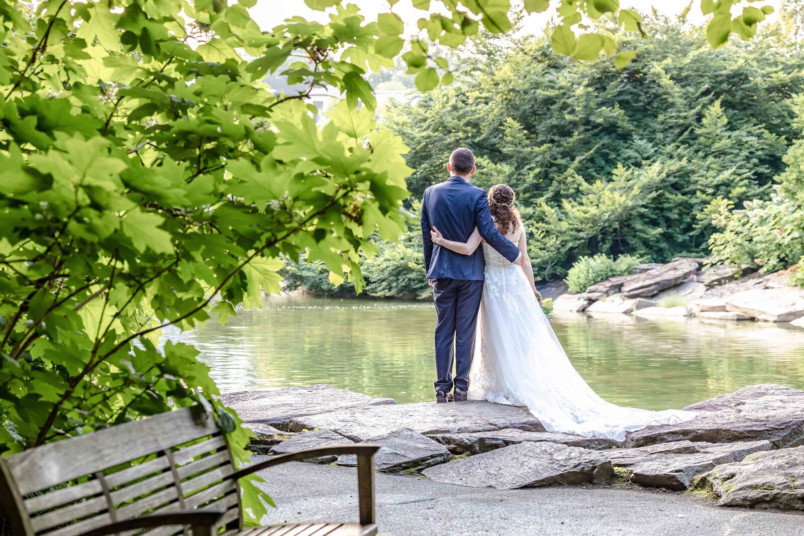 The bride and groom stand arm-in-arm by a tranquil lakeside surrounded by lush greenery, captured during an Asheville, NC wedding, blending rustic elegance with serene natural beauty.