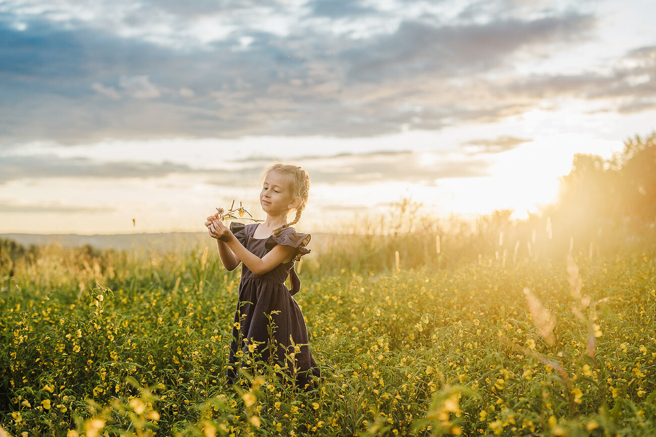 girl picks flowers in field of yellow flowers with dramatic clouds and sunset