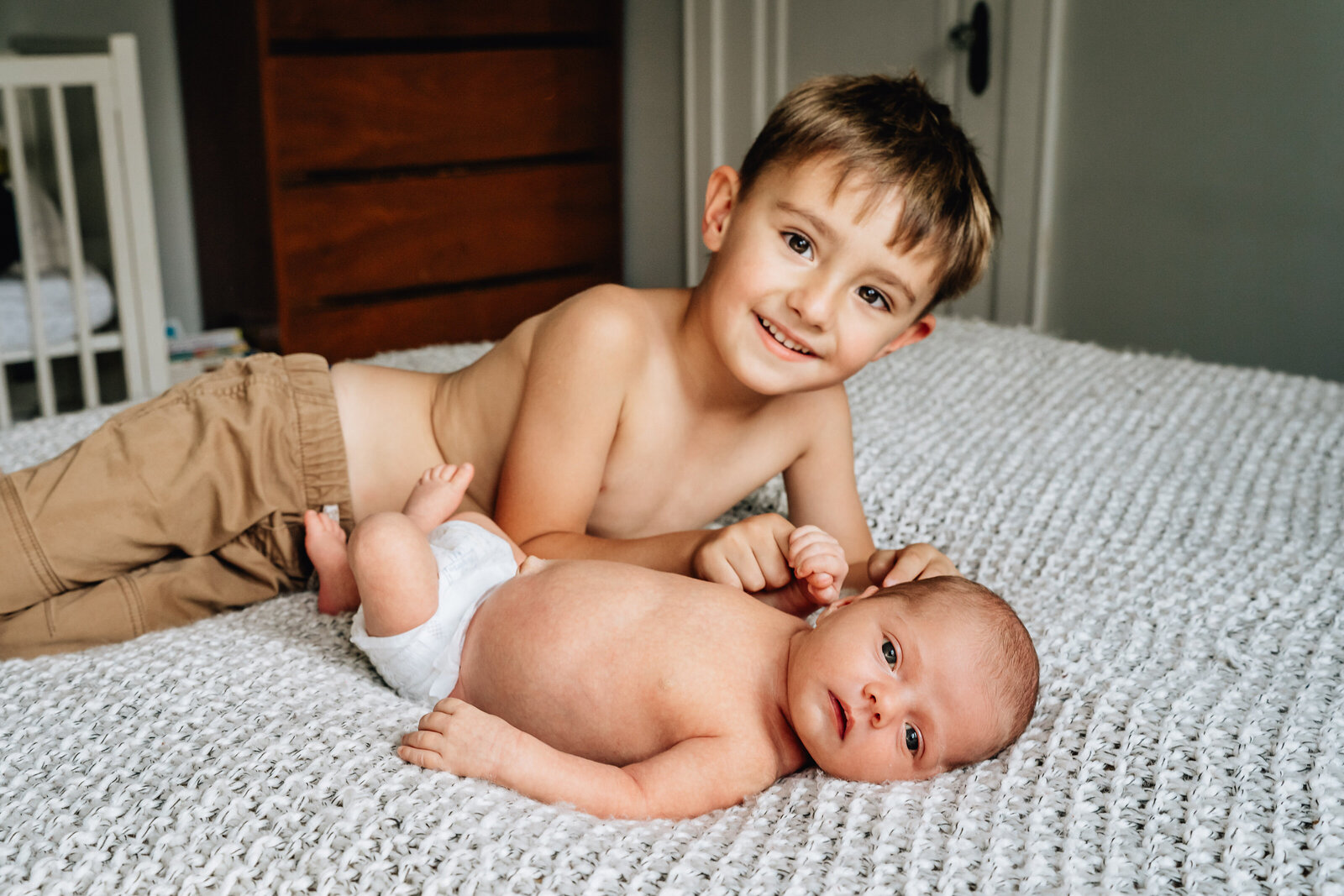 A big brother lies on a bed next to his newborn sibling, both with warm expressions.