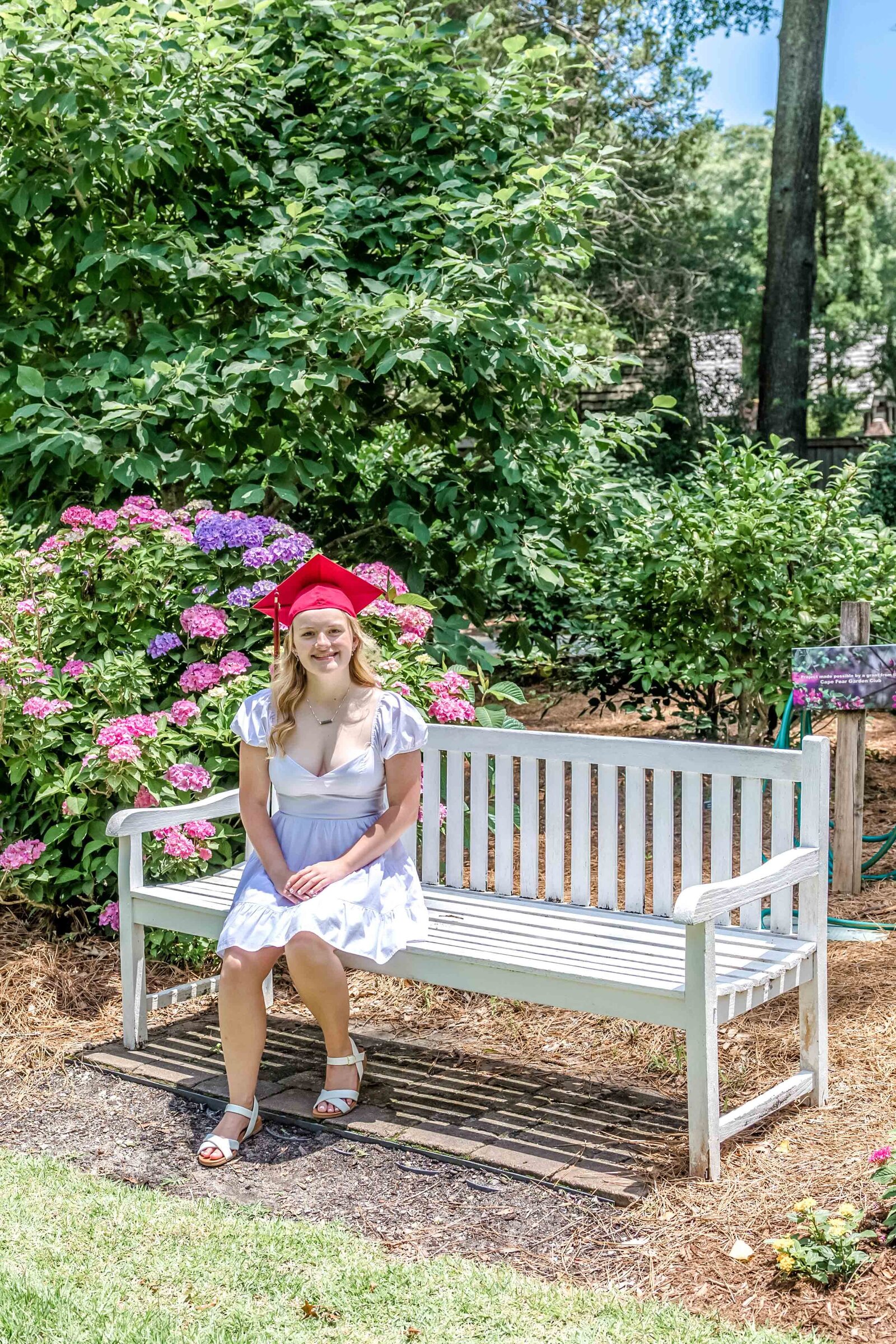 A high school senior in a red cap sitting on a white bench surrounded by blooming flowers in North Carolina. This charming scene captures the beauty of outdoor senior photography, perfect for those wanting vibrant and memorable graduation photos.
