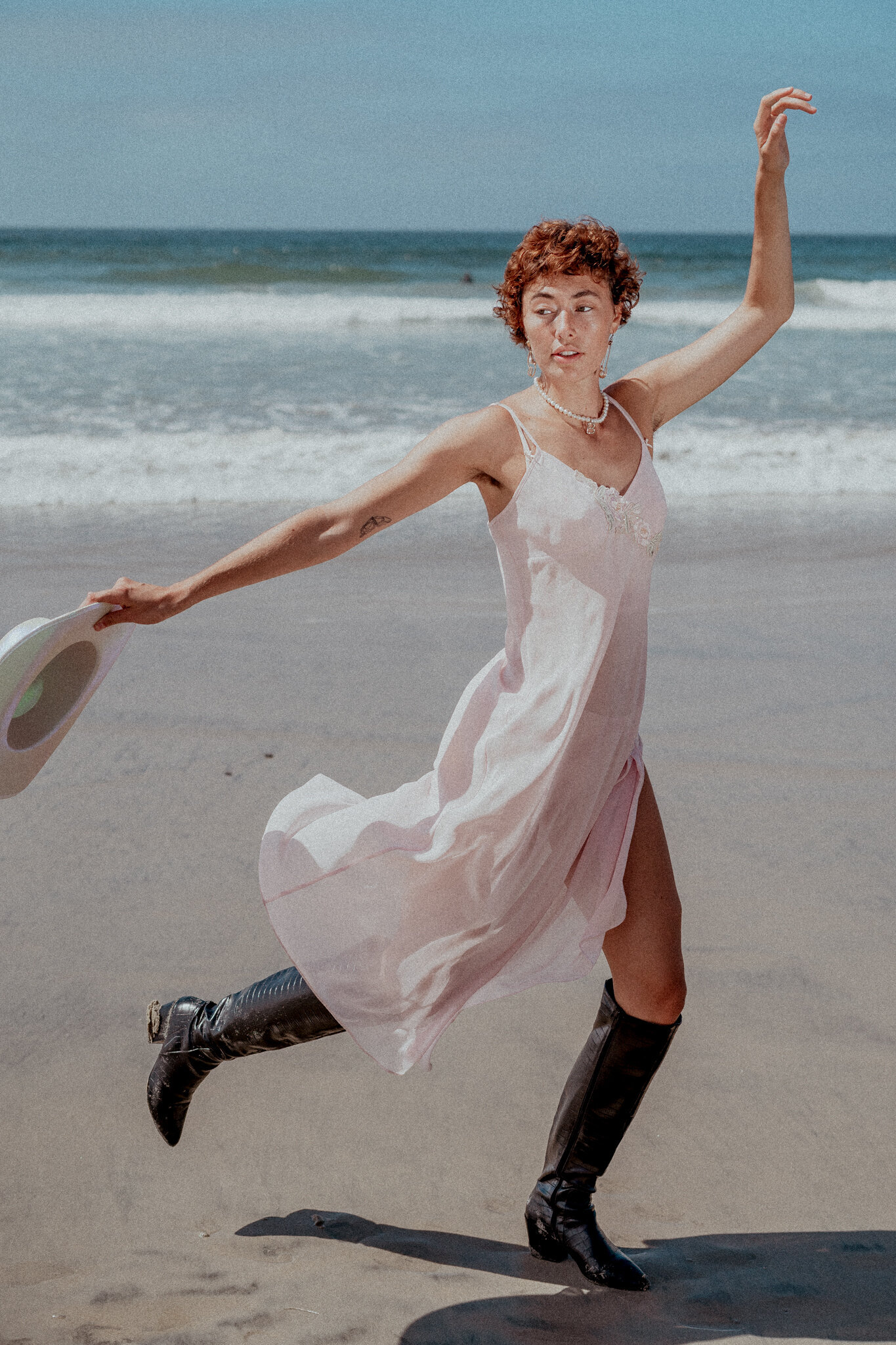 Young woman wearing a silk pink dress and black cowboy boots twirling at the beach