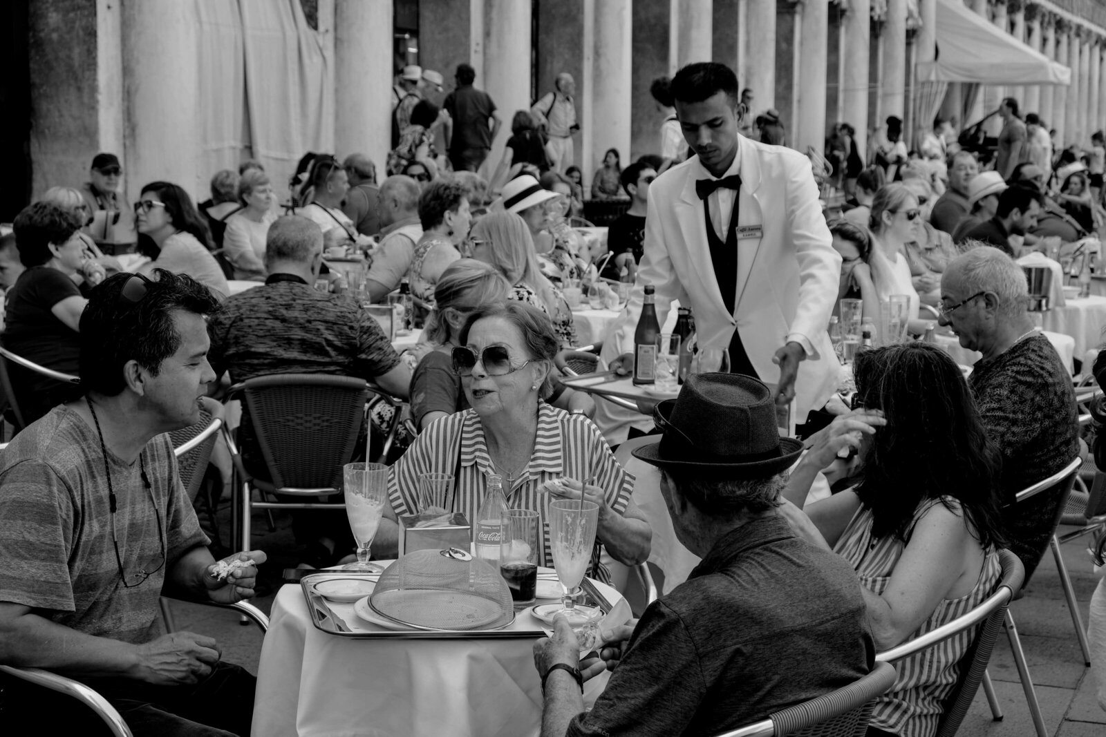 Waiter in white coat and black bow tie brings food and drinks to diners sitting at caffe in Piazza San Marco