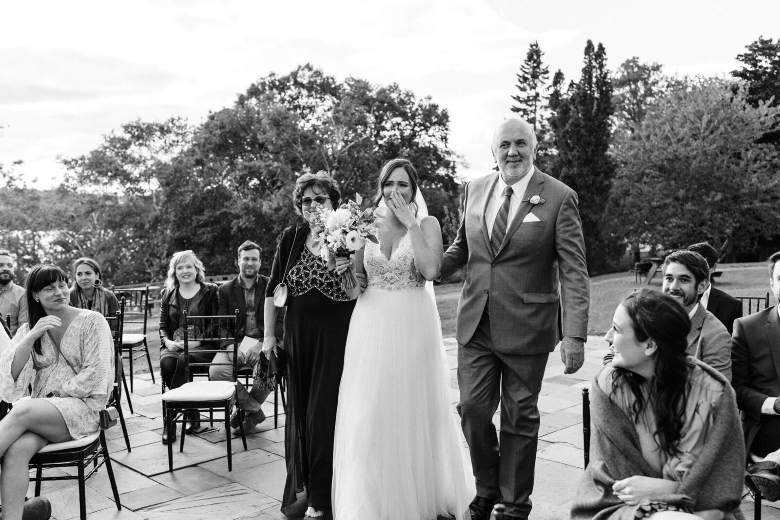 Candid moment of bride walking down aisle with parents at outdoor wedding ceremony.