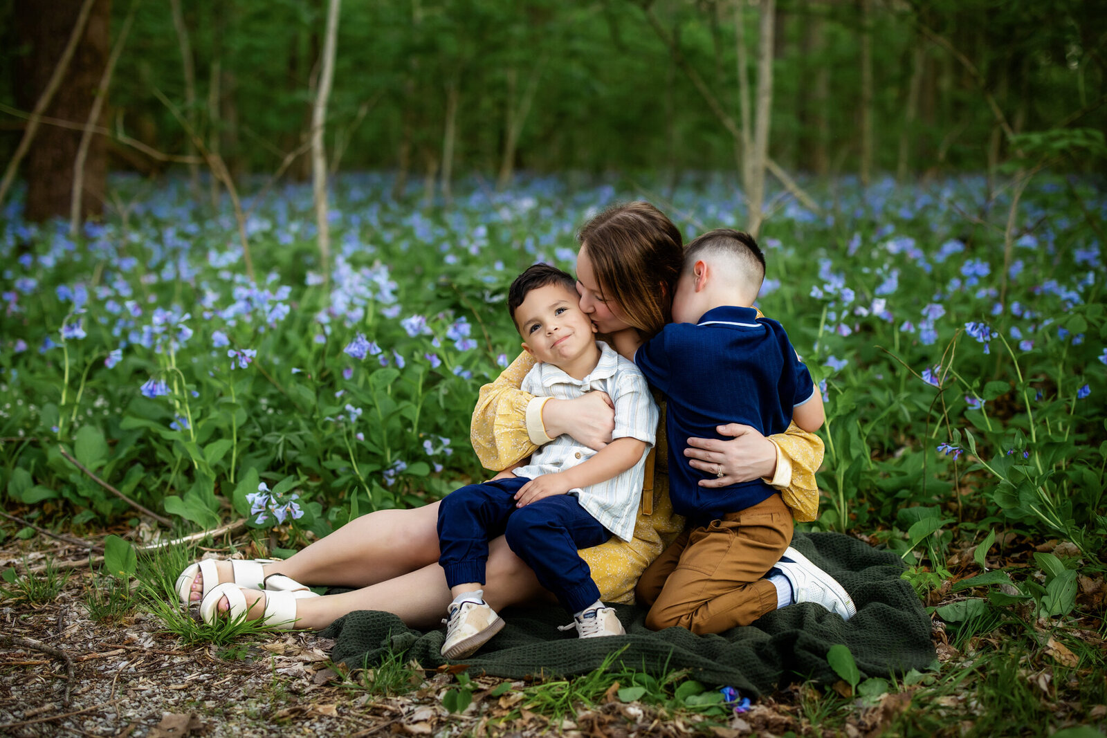 A woman sits on a blanket in a field of blue flowers, hugging two young boys. Captured by a Bloomington family photographer, the outdoor portrait radiates warmth as one boy smiles at the camera while the other hugs her and rests his head on her shoulder.