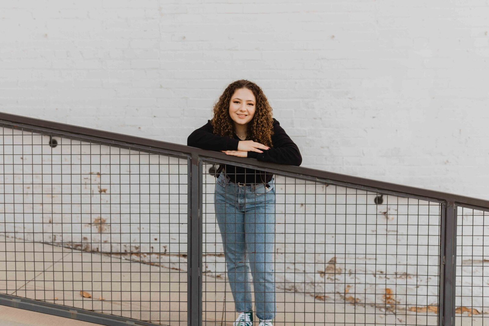 Senior girl standing in front of white wall, leaning on railing with arms rested on it. Smiling and looking at the camera.