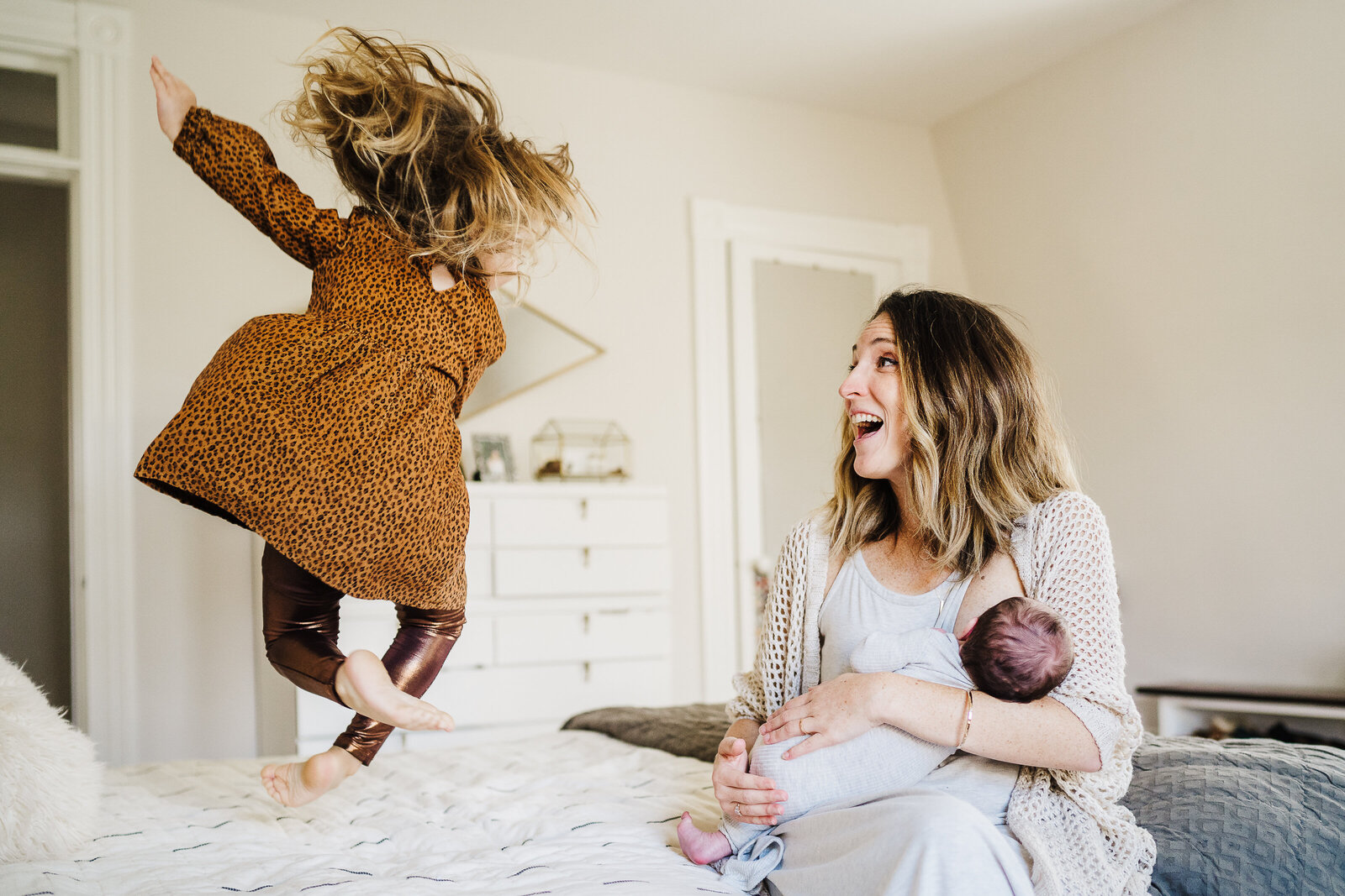 mother holding newborn laughs as toddler jumps on bed