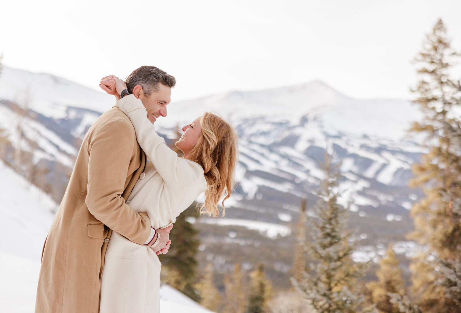A husband and wife are facing one another, wrapped in each others arms on a snowy day. Breckenridge resort with all the ski runs can be seen behind them, 
