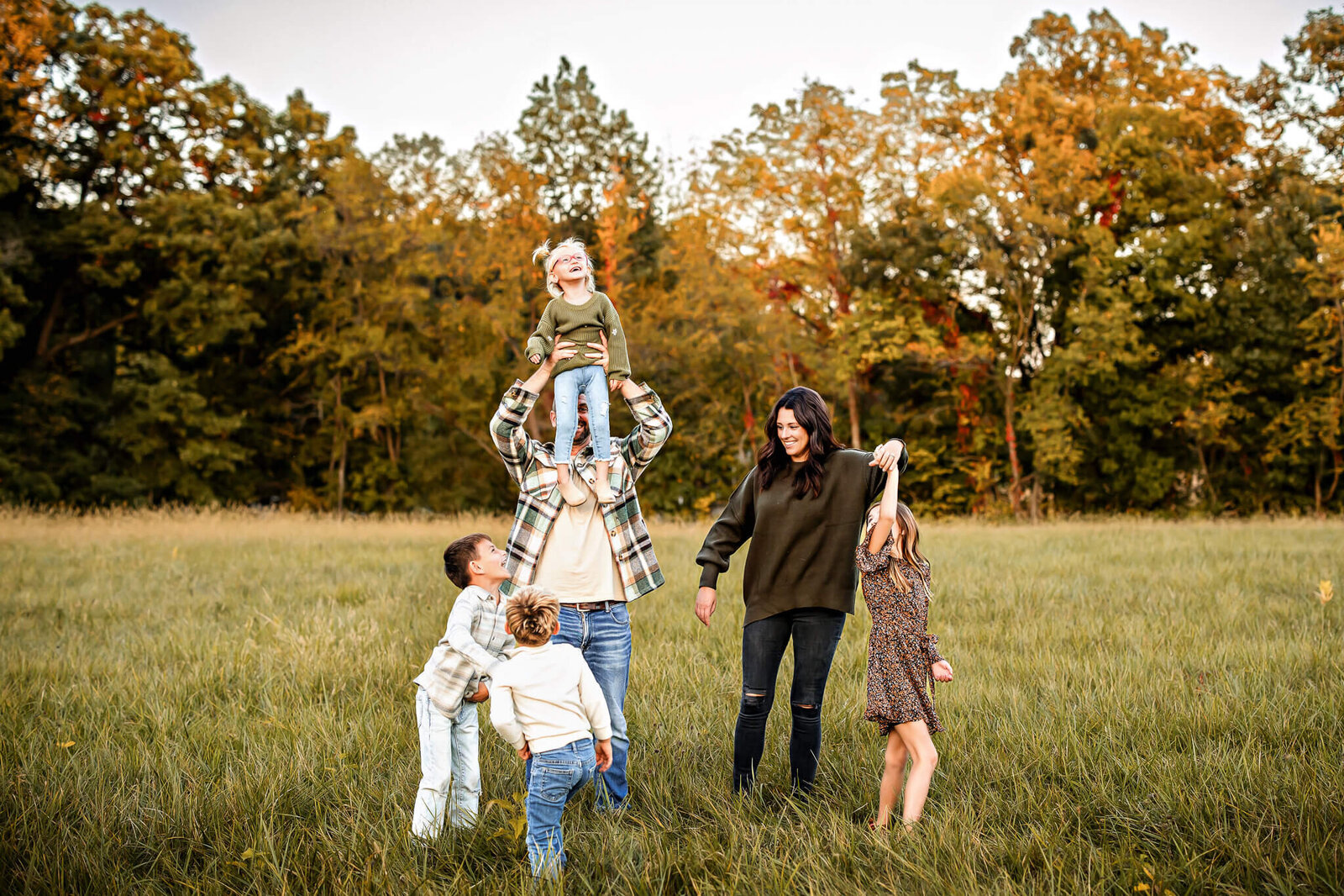 A family of six enjoys a day in a grassy field with autumn trees as their backdrop. A man lifts a child onto his shoulders, while a woman and three other children play nearby. Everyone is laughing and wearing casual fall clothing, embodying the beauty of outdoor portraits in Bloomington.