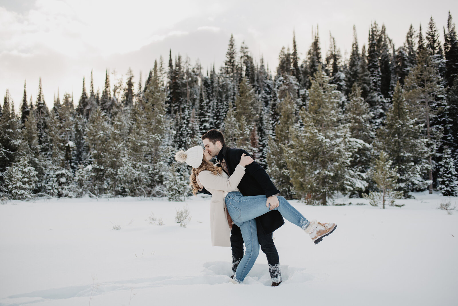 grand tetons engagement pictures with man dipping woman backwards while kissing passionately in front of a wooded area in a snowy field captured by jackson hole photographers