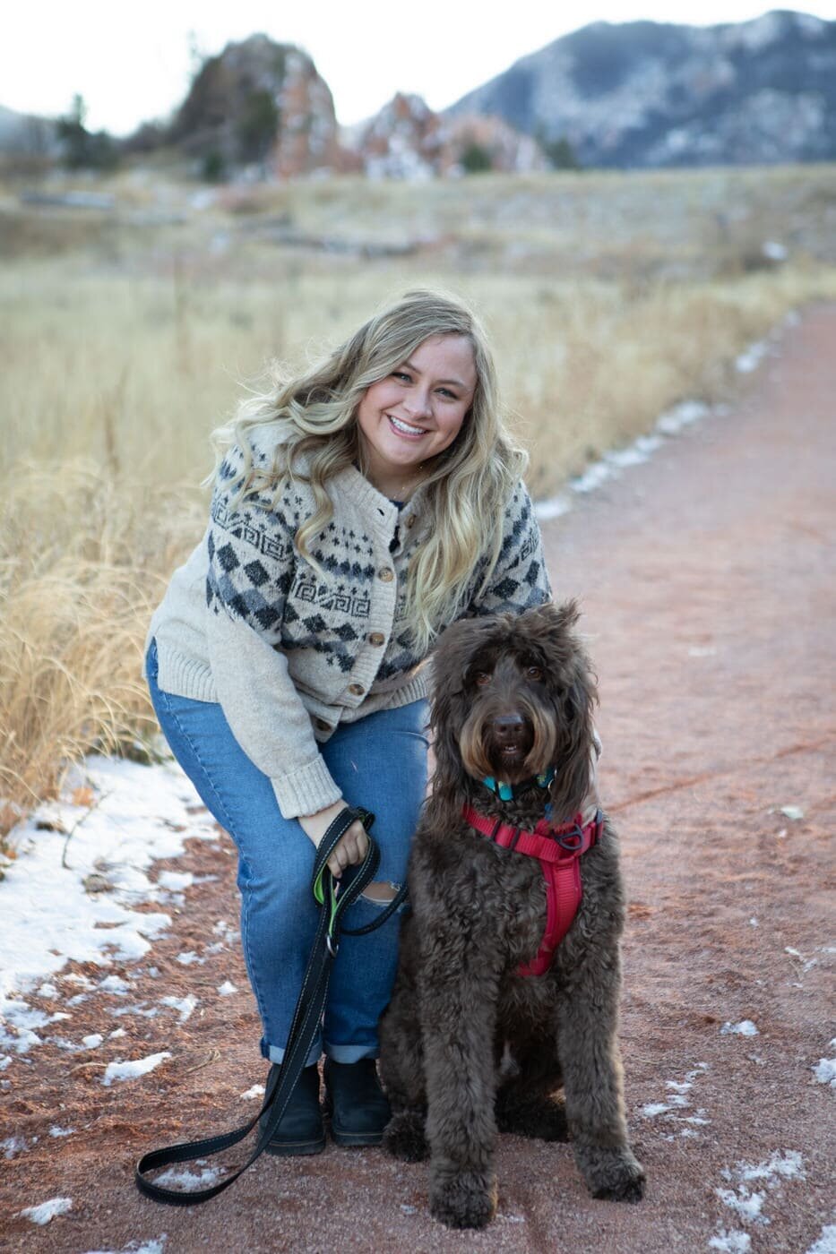 A woman crouches down towards her labradoodle  as they both look at the camera.