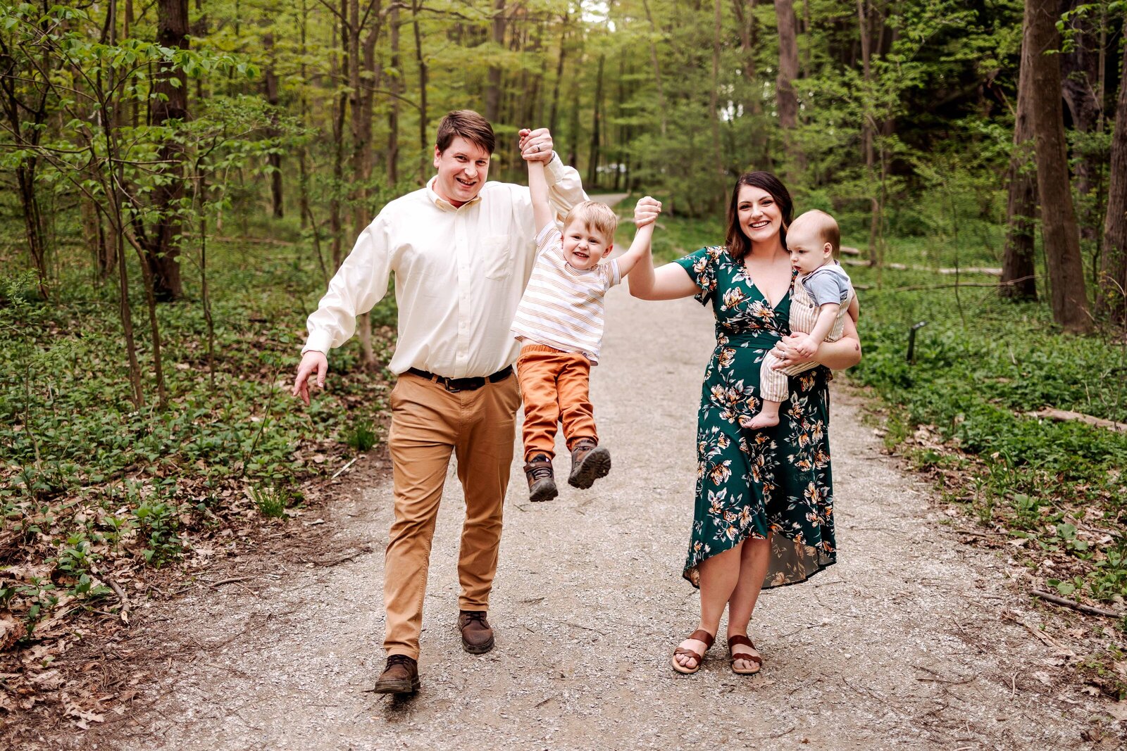 Family swing their toddler forward for a fun photo along a wooded path