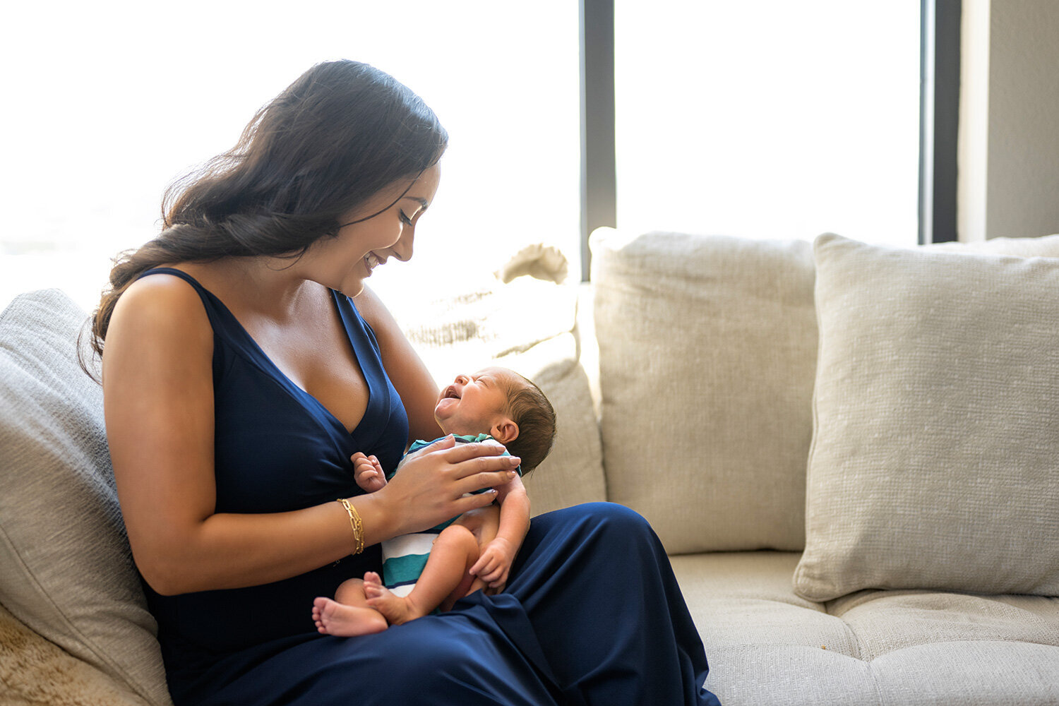 A mother smiles down at her newborn in natural window light during an in-home Dallas session