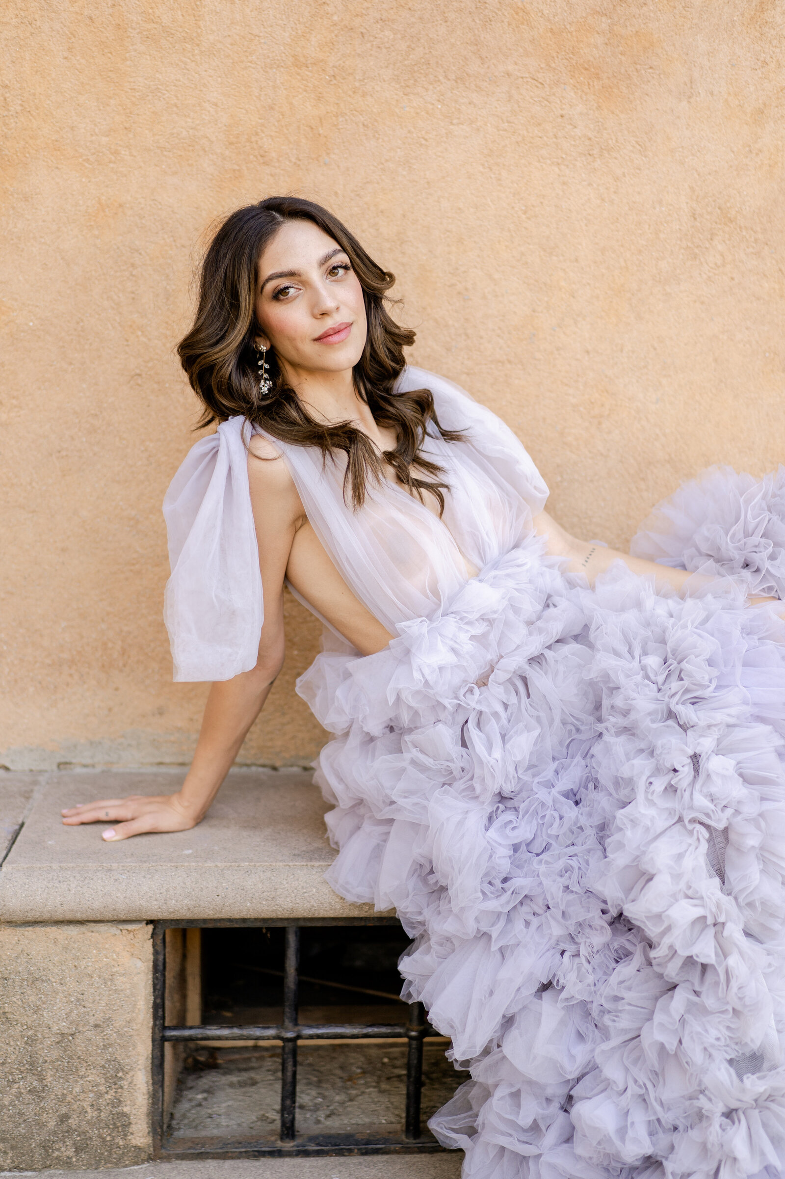 Portrait of a bride in a lavender gown sitting on a concrete stoop in front of a peach wall.