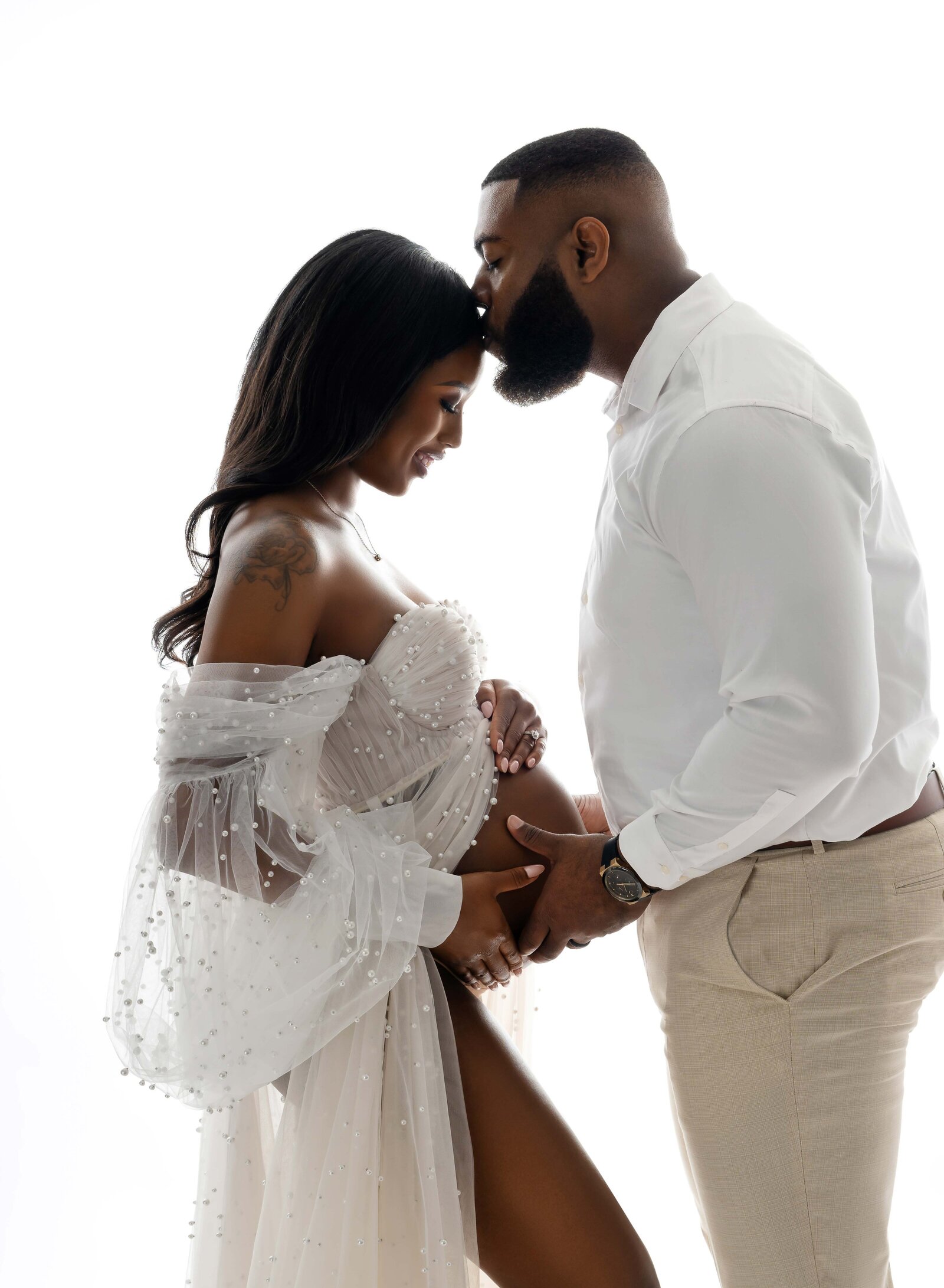 Male, holding baby bump and kissing pregnant women's head. Pregnancy photo taken in studio on white backdrop.
