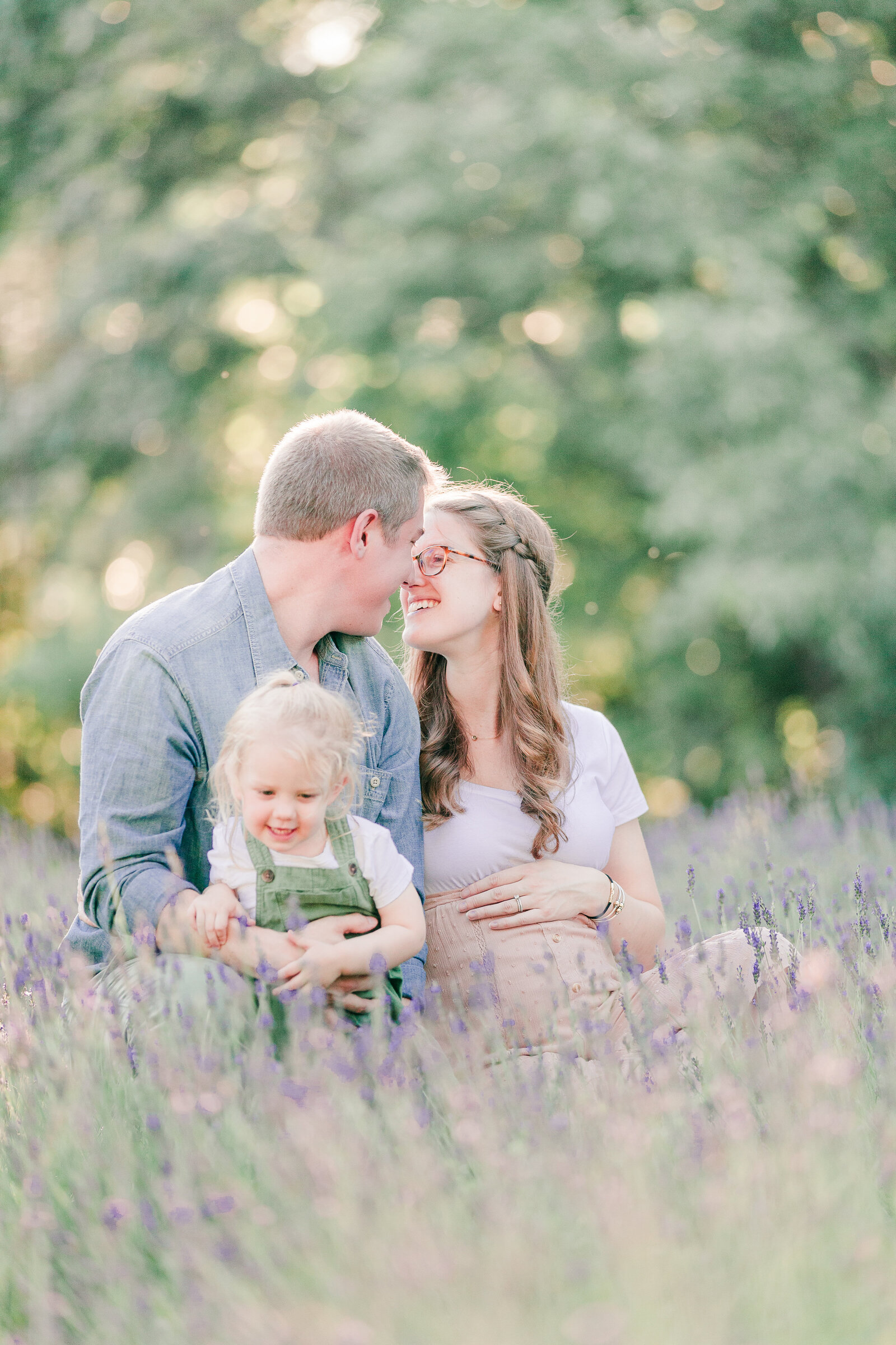 Pregnant mom with hand gently on her baby bump, sitting with little girl and dad in a lavender field by NH newborn photographer Fieldstone Studio.