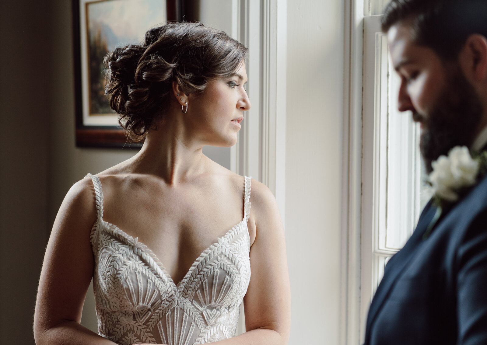 A bride in a vintage dress looks out a large window