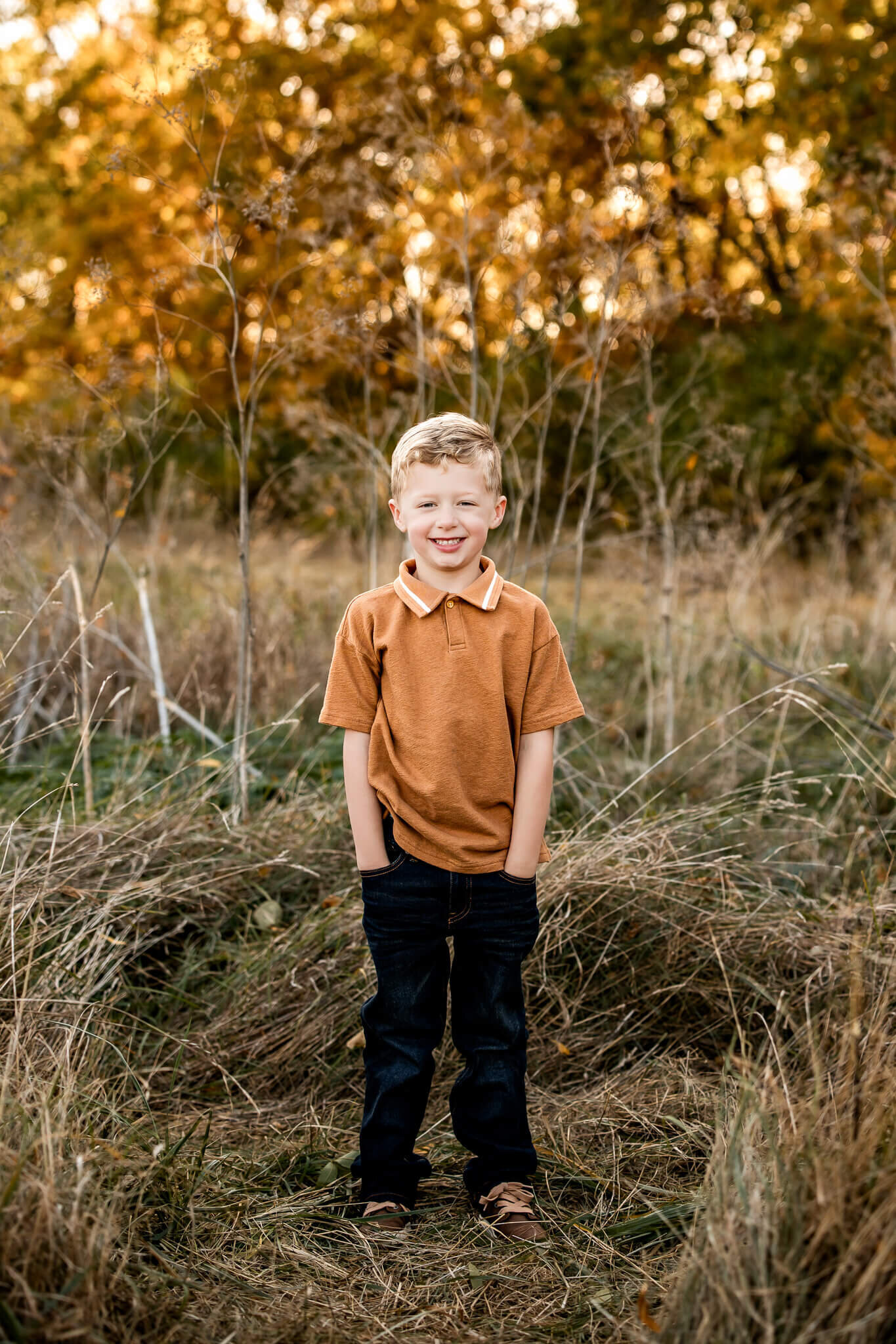 A young boy stands smiling with his hands in his pockets in a field, capturing the beauty of outdoor portraits. He is wearing a brown shirt and dark jeans. Tall grass and autumn trees with golden leaves form the background, reminiscent of a Bloomington family photographer's touch.