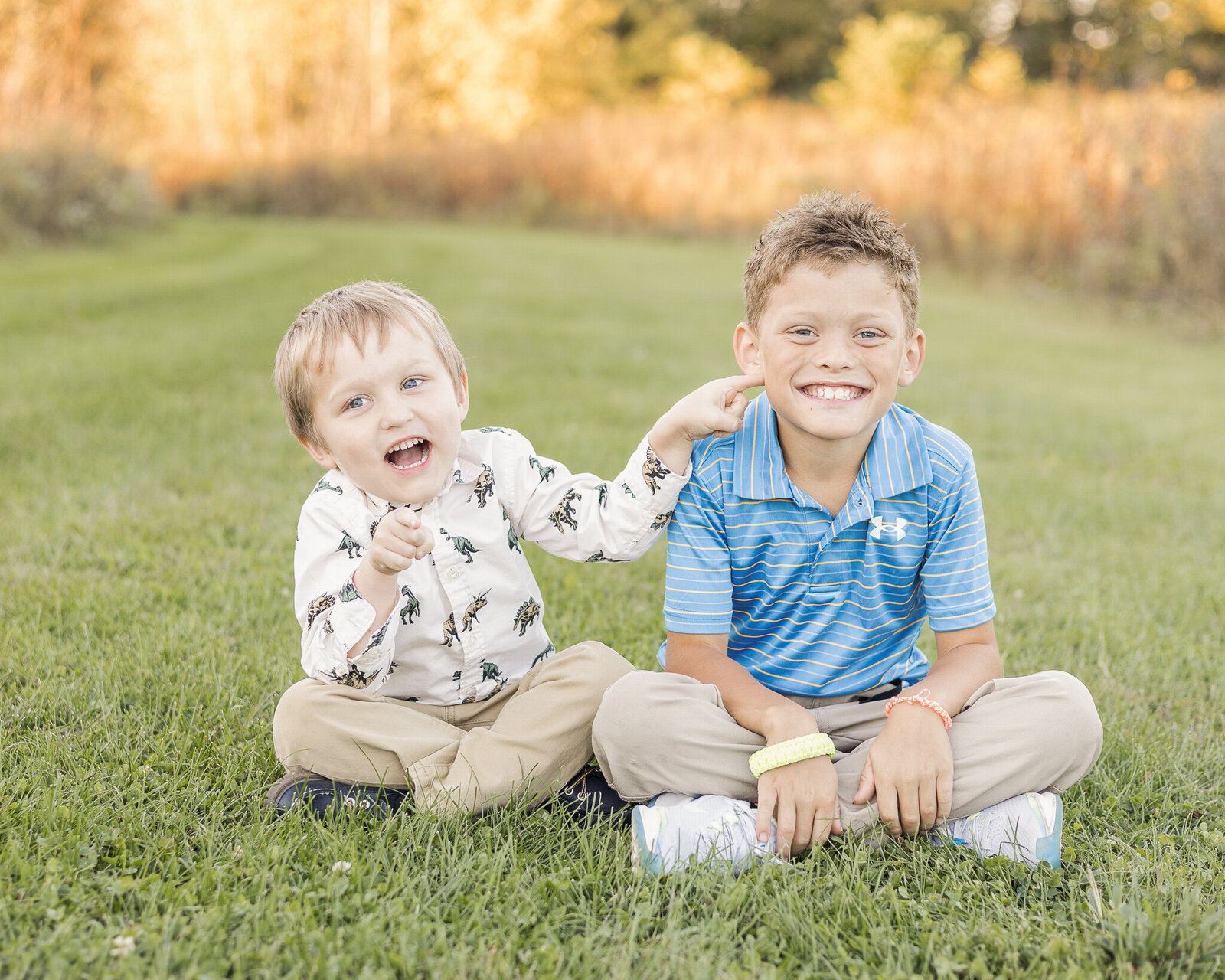 Two young boys sitting in the grass in the park.  One has light brown blonde hair, wearing a white print shirt, khaki pants, and smiling away from the camera.  The other boy has sandy blonde hair, wearing a blue striped shirt, khaki pants and smiling at the camera