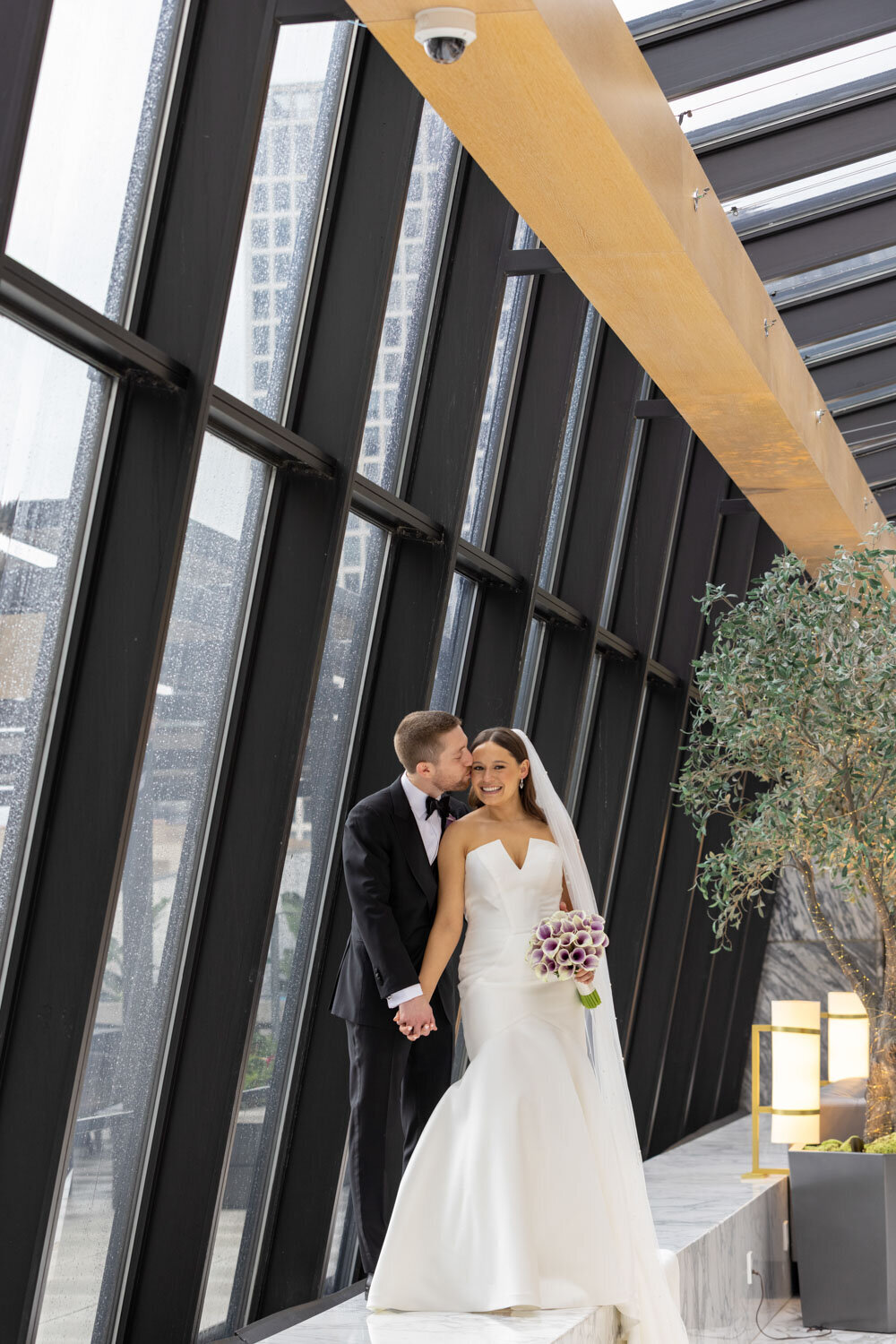 A bride and groom kissing in front of a large window