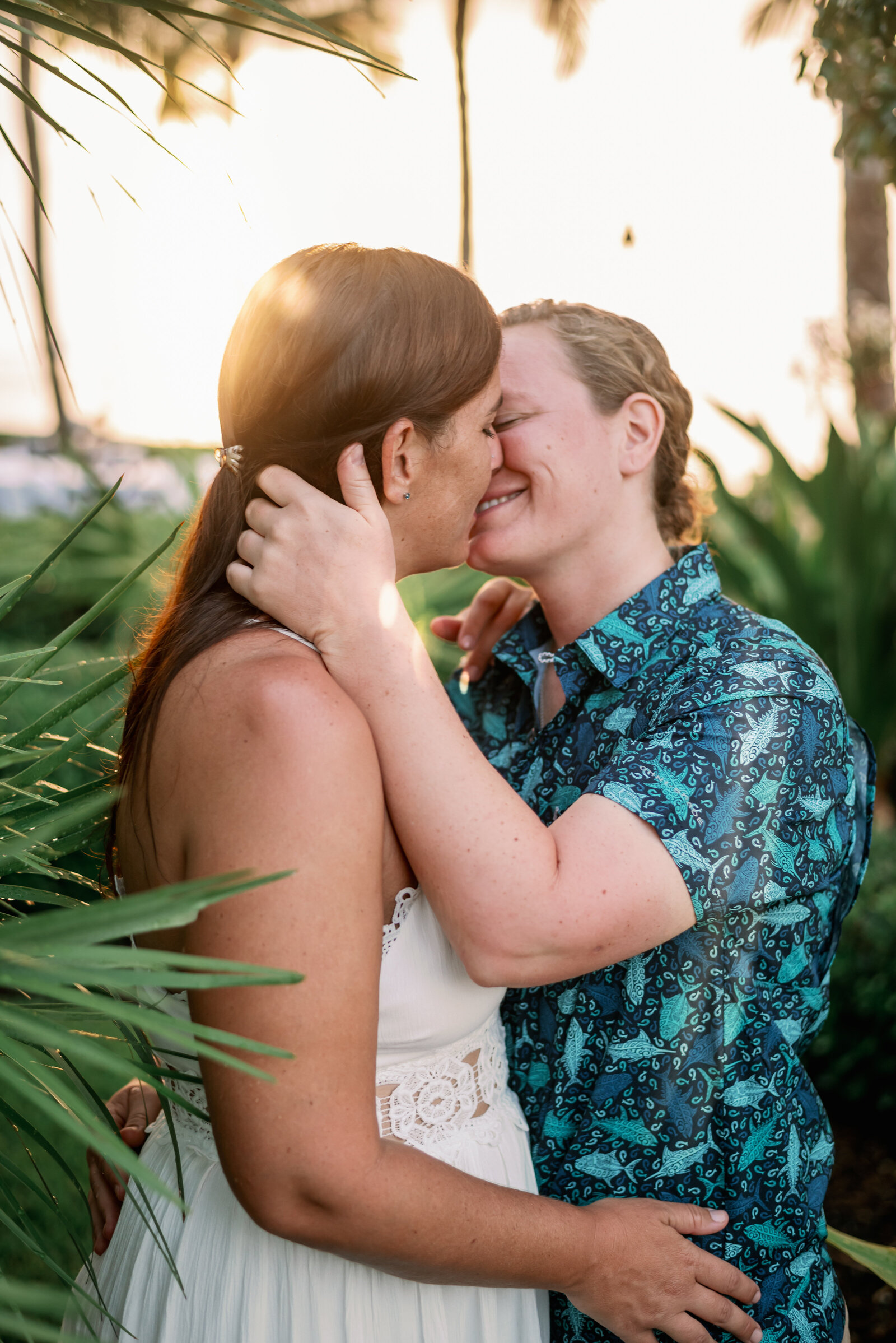LGBTQ+ engaged couple giving a tender kiss while the sun sets behind them