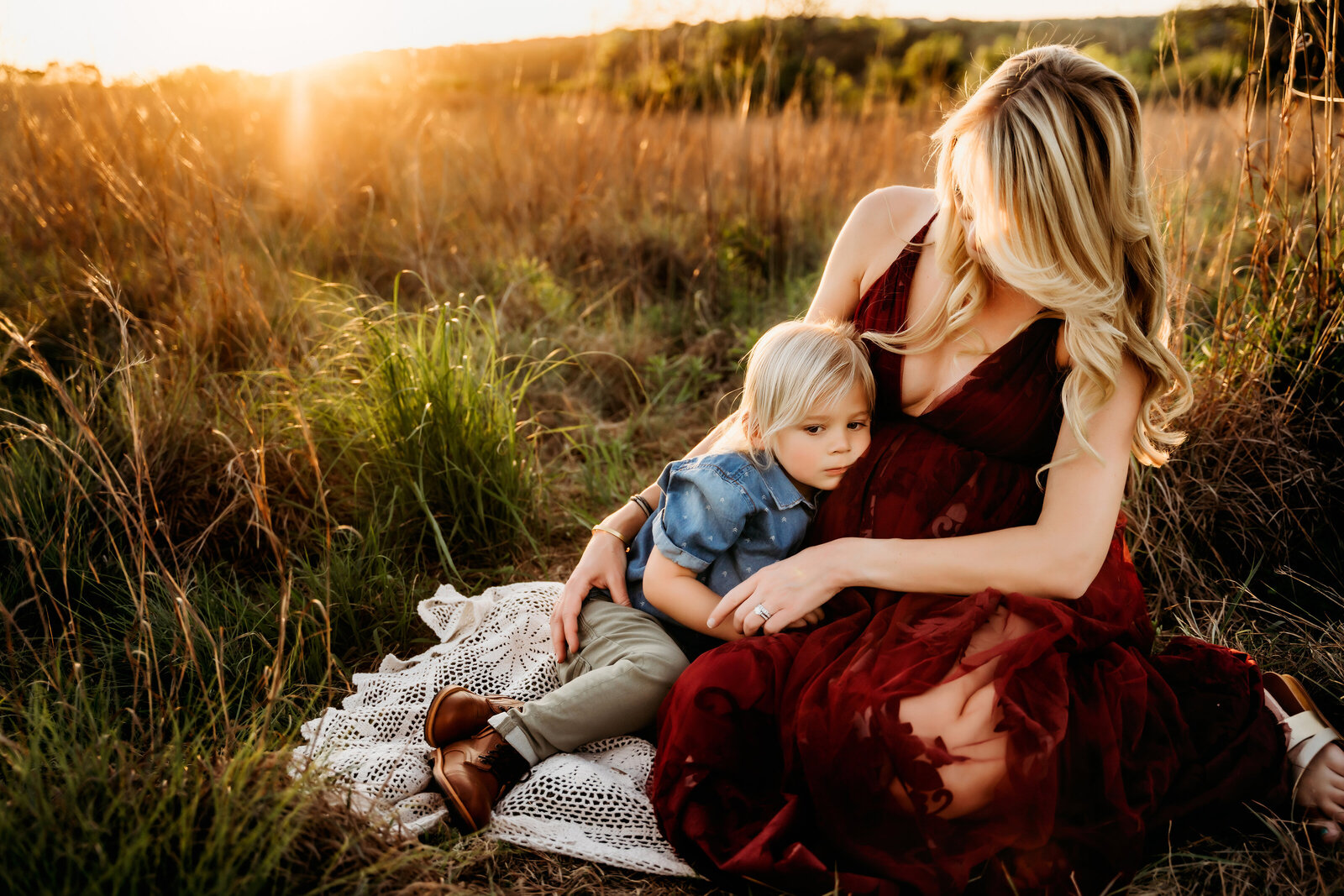 Maternity Photographer, a pregnant mother holds her young son as they sit in tall grass at golden hour