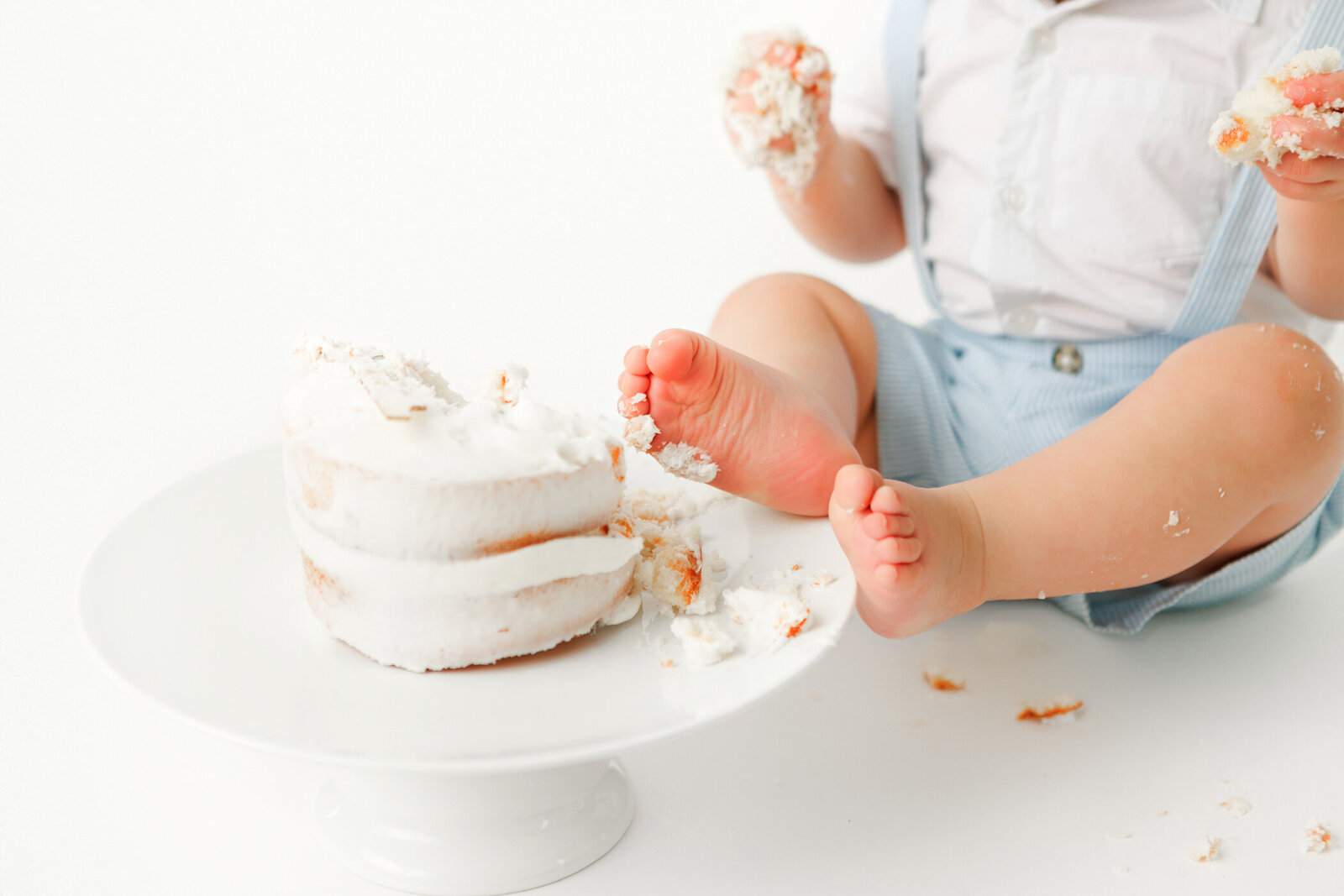 First Birthday cake smash photo session in fresno, ca. all white, neutral, simple style cake with baby boy dressed in blue romper with his feet on the cake plate