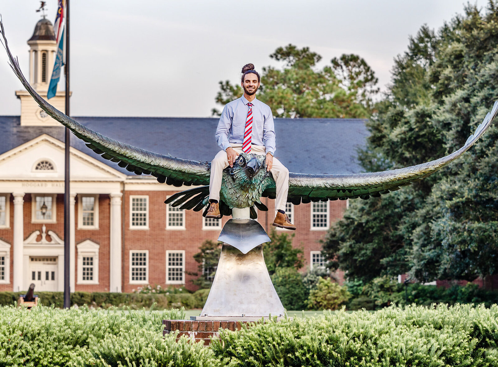 A college senior in a tie sitting atop the seahawk sculpture on the University of North Carolina campus in Wilmington, North Carolina. This unique photo showcases individuality and achievement, perfect for those seeking creative and professional senior photography.