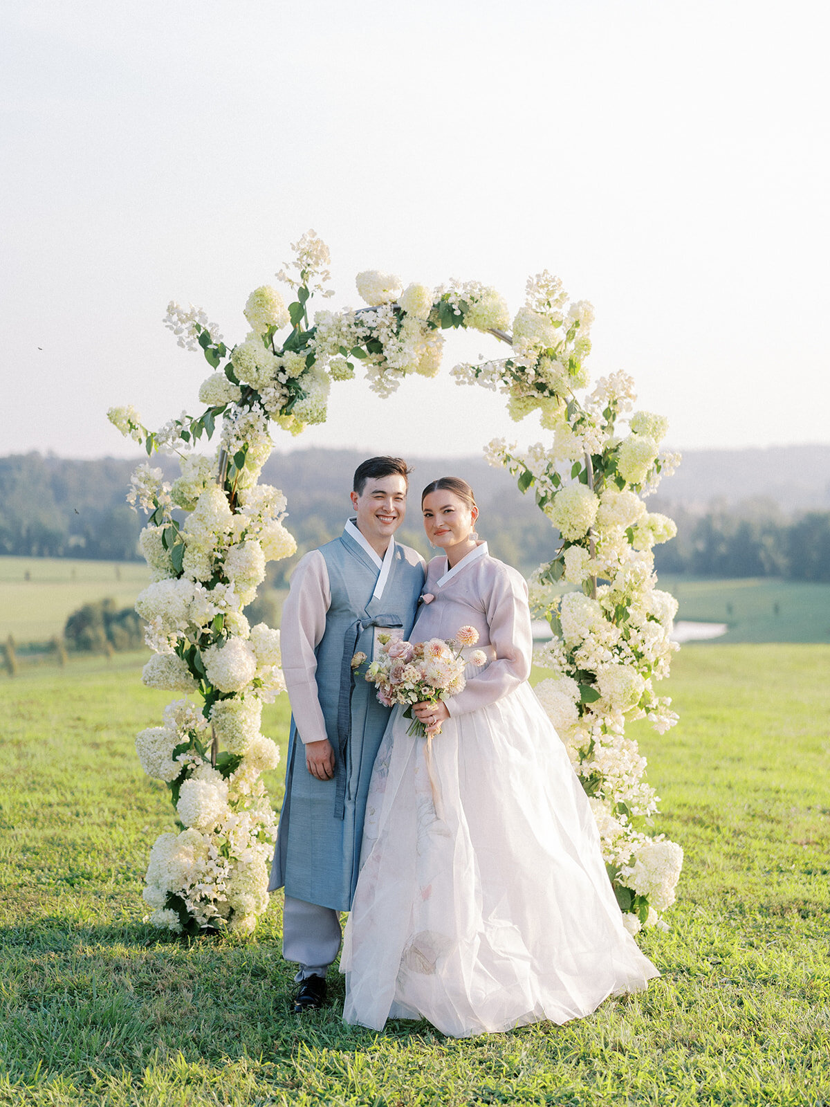 Portrait of bride and groom in traditional Korean hanboks under a full floral arch filled with white, airy hydrangea.