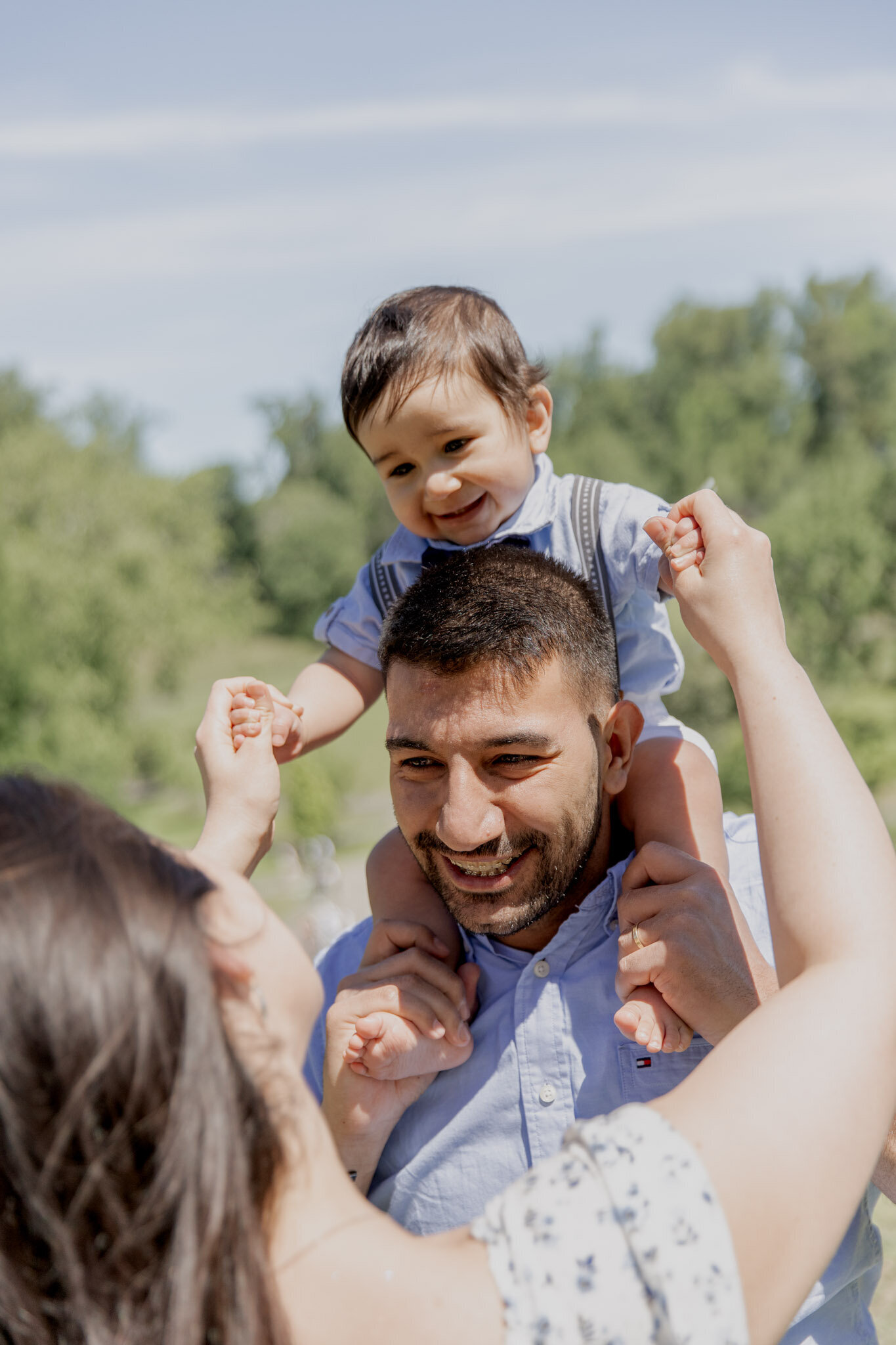 Baby boy is sitting on dad's shoulders, both him and dad are smiling down at mom, who is facing him and holding his hands.