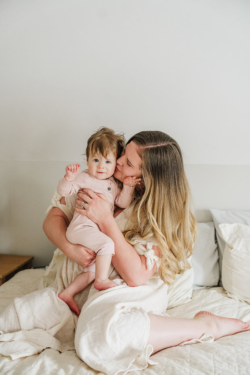 mom in white dress snuggles daughter on white bed