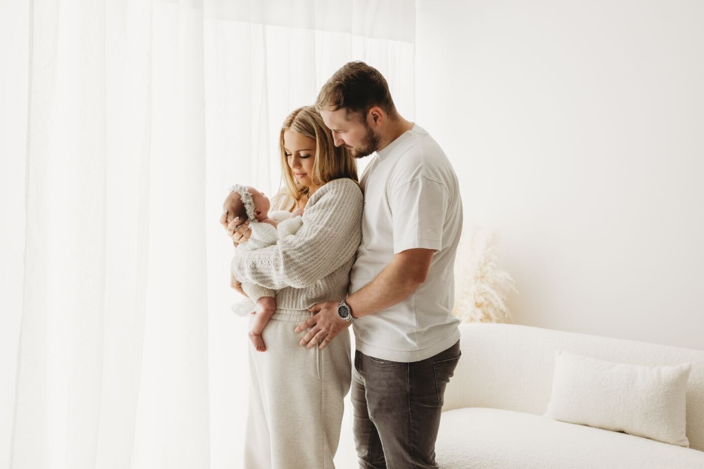 a mother and father lovingly holding their peacefully sleeping newborn baby near a bright window by Ruaa Hamdan Photography in UK