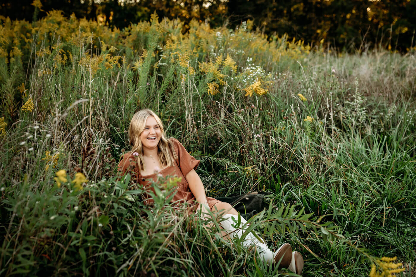 A person with long hair, wearing a brown dress, is sitting and smiling amidst tall grass and yellow wildflowers, with a background of trees under a clear sky.