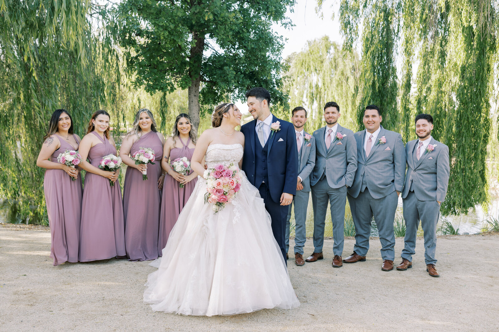 bridal party pictures by sacramento wedding photographer with bride and groom standing together and looking at one another and smiling with their bridal party standing behind them posing and smiling with the lawn behind them with weeping willow trees