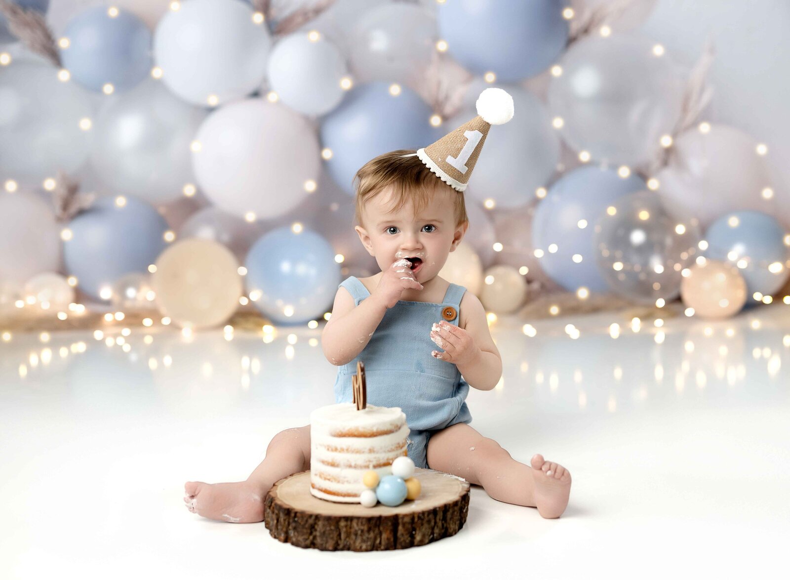 Baby boy in blue romper eating cake in front of white and blue balloon garland with lights. Photographed by Jenni Nikki at Atlanta Photo studio.