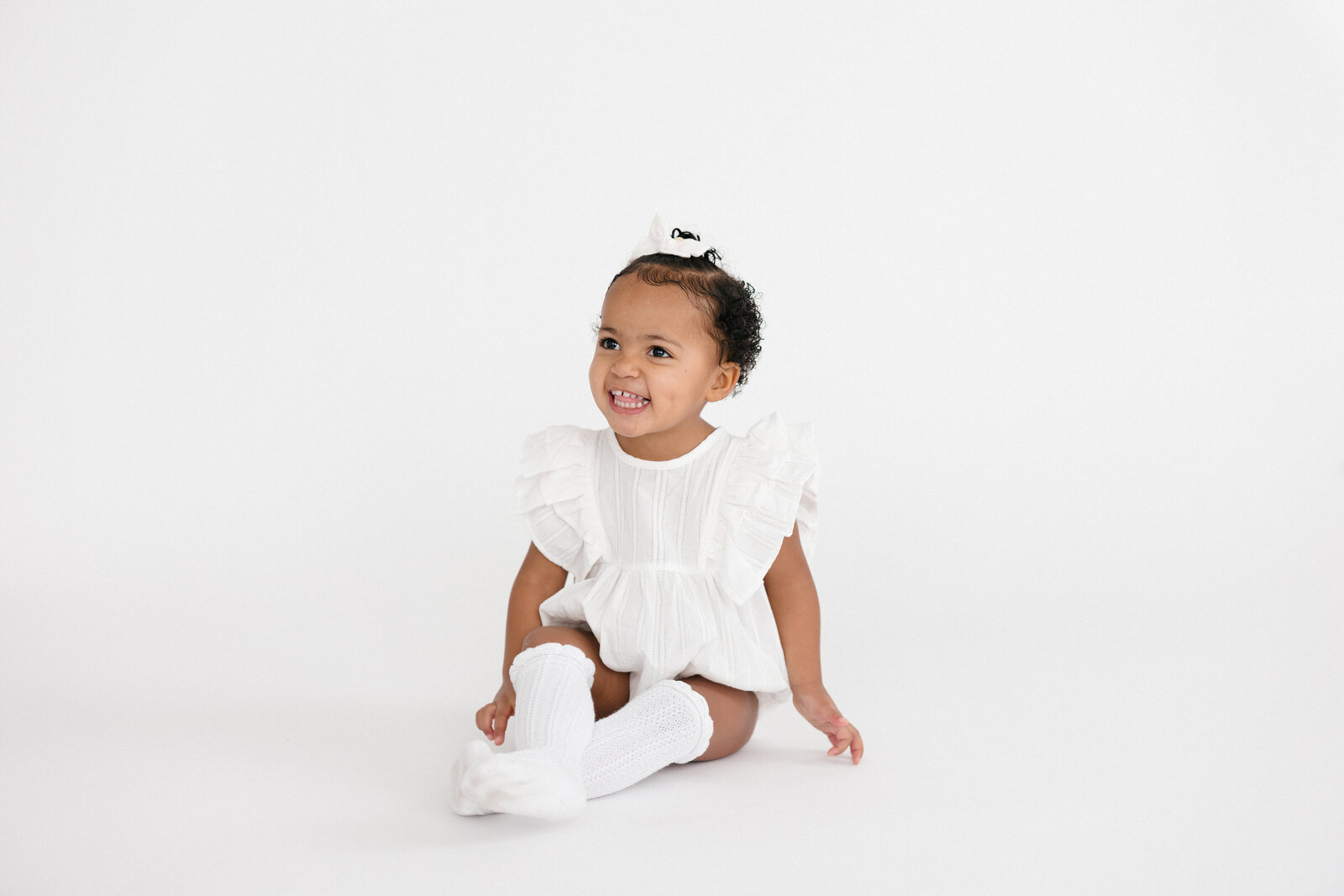 Baby girl sitting on a white backdrop wearing a white outfit and white knee-high socks smiling and laughing
