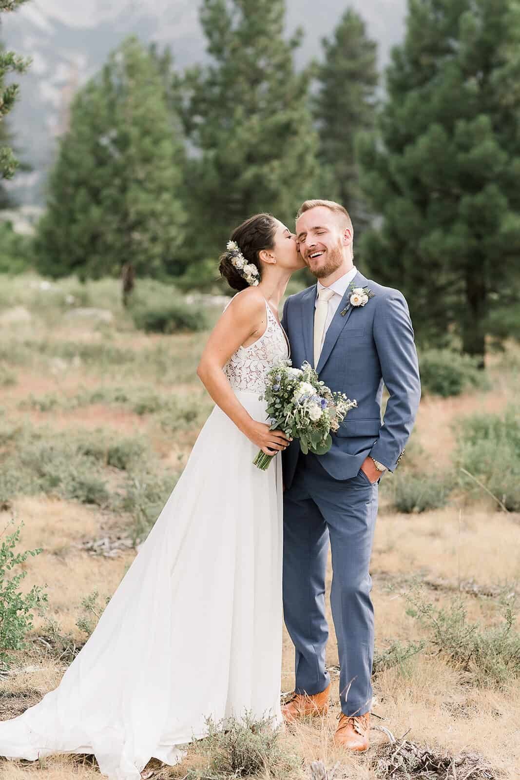 A bride kisses her groom on the cheek in front of pine trees while on their Mammoth wedding day. 