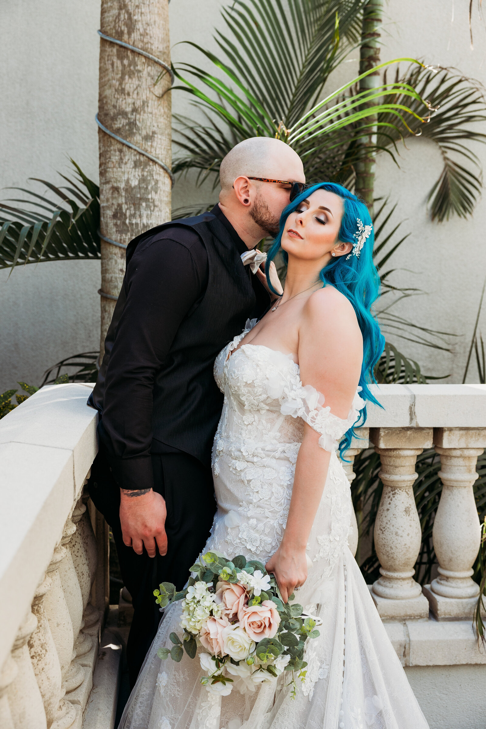 Groom kisses bride under palm trees on a balcony in downtown St. Augustine, her bouquet filled with blush roses and white florals.