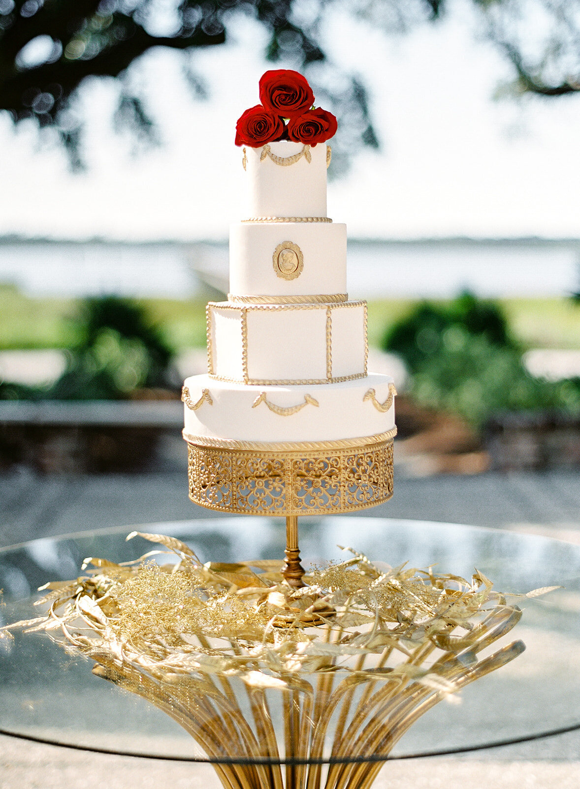 Ivory and gold four tier wedding cake on gold pedestal with painted gold myrtle leaves around the glass top of the table with gold table legs. Photographed at Lowndes Grove wedding reception by wedding photographers in Charleston Amy Mulder Photography.
