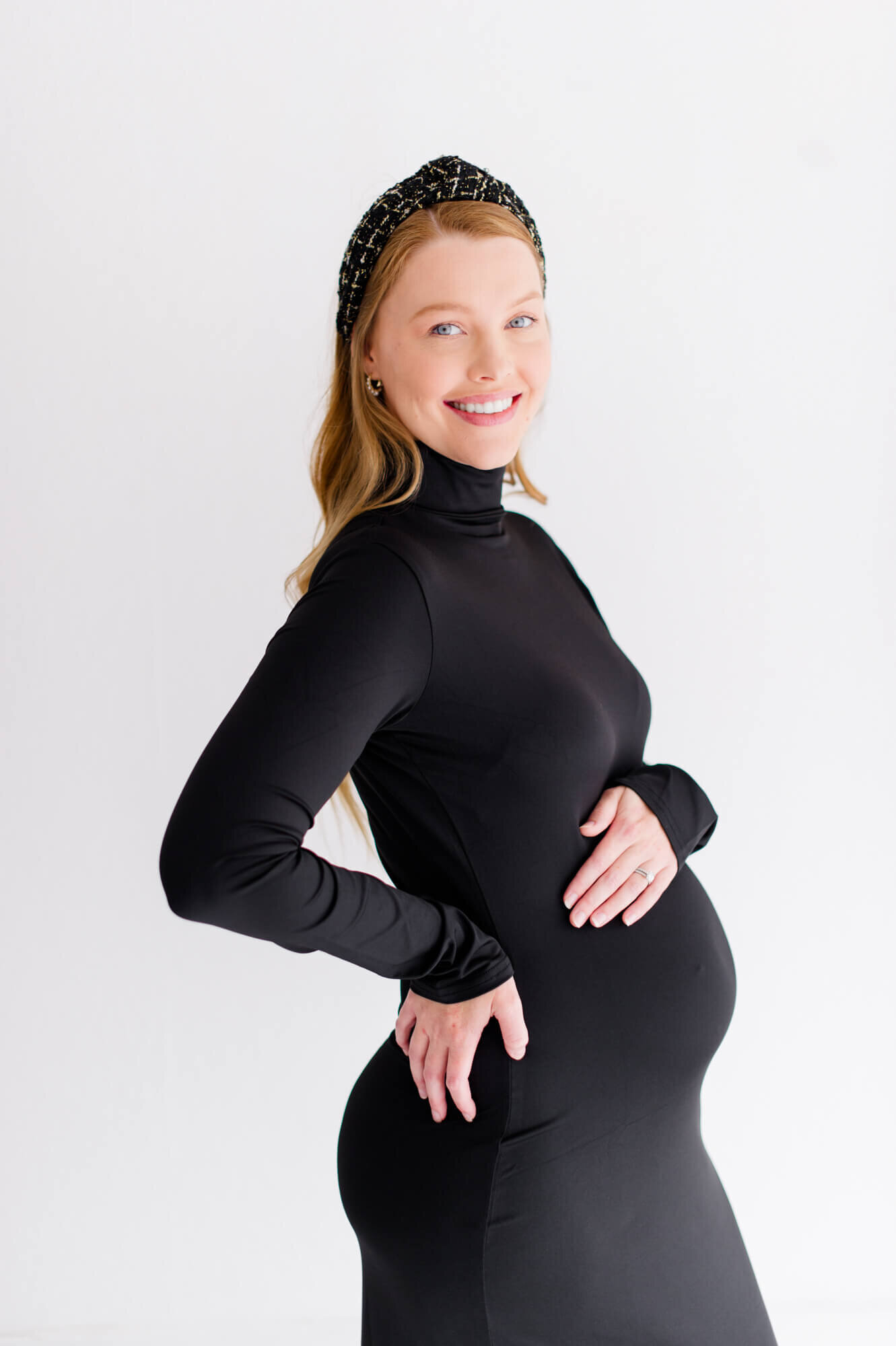 Pregnant mom stands holding belly and smiling at camera during her studio maternity session