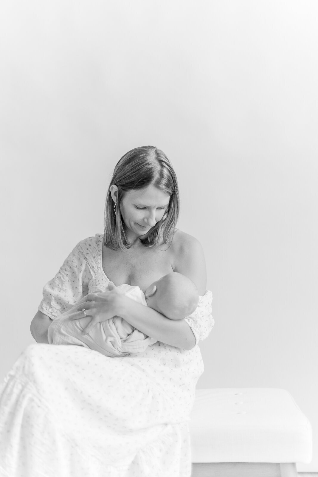 A black and white photo of a young mother looking down at her baby boy while she breastfeeds him while wearing an off the shoulder dress during photo session with Boston family photographer Corinne Isabelle