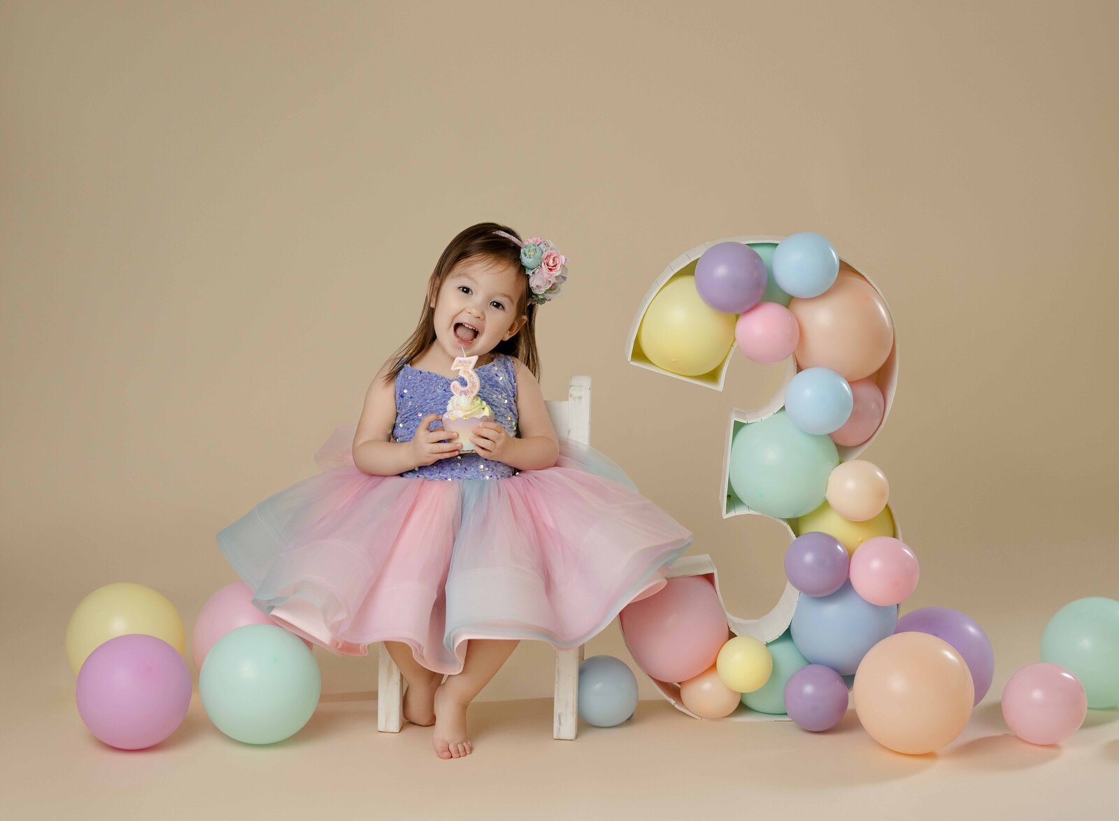 3rd birthday photo of girl in pastel tulle dress with cupcake. Surprise expression on her face and surrounded by pastel balloons. Photographed by Atlanta birthday Photographer.