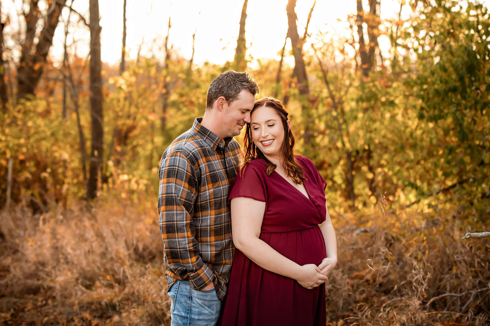 A couple stands close in a sunlit forest, autumn foliage around them. The woman in a burgundy dress smiles down as the man in plaid leans towards her—capturing a tender moment perfect for maternity portraits or pregnancy photography.