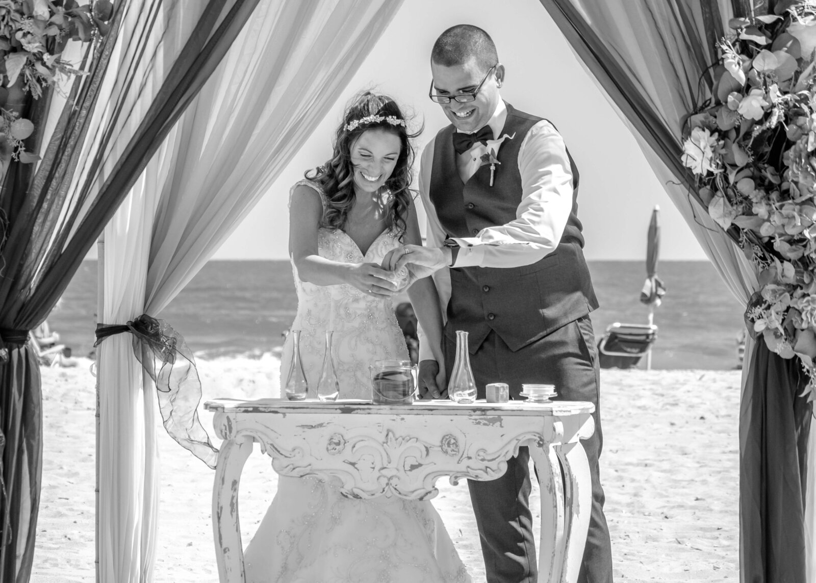 The bride and groom participate in a sand ceremony under a draped beach arbor, with the ocean in the background, captured during a romantic Topsail Beach, NC wedding, blending tradition and coastal charm.