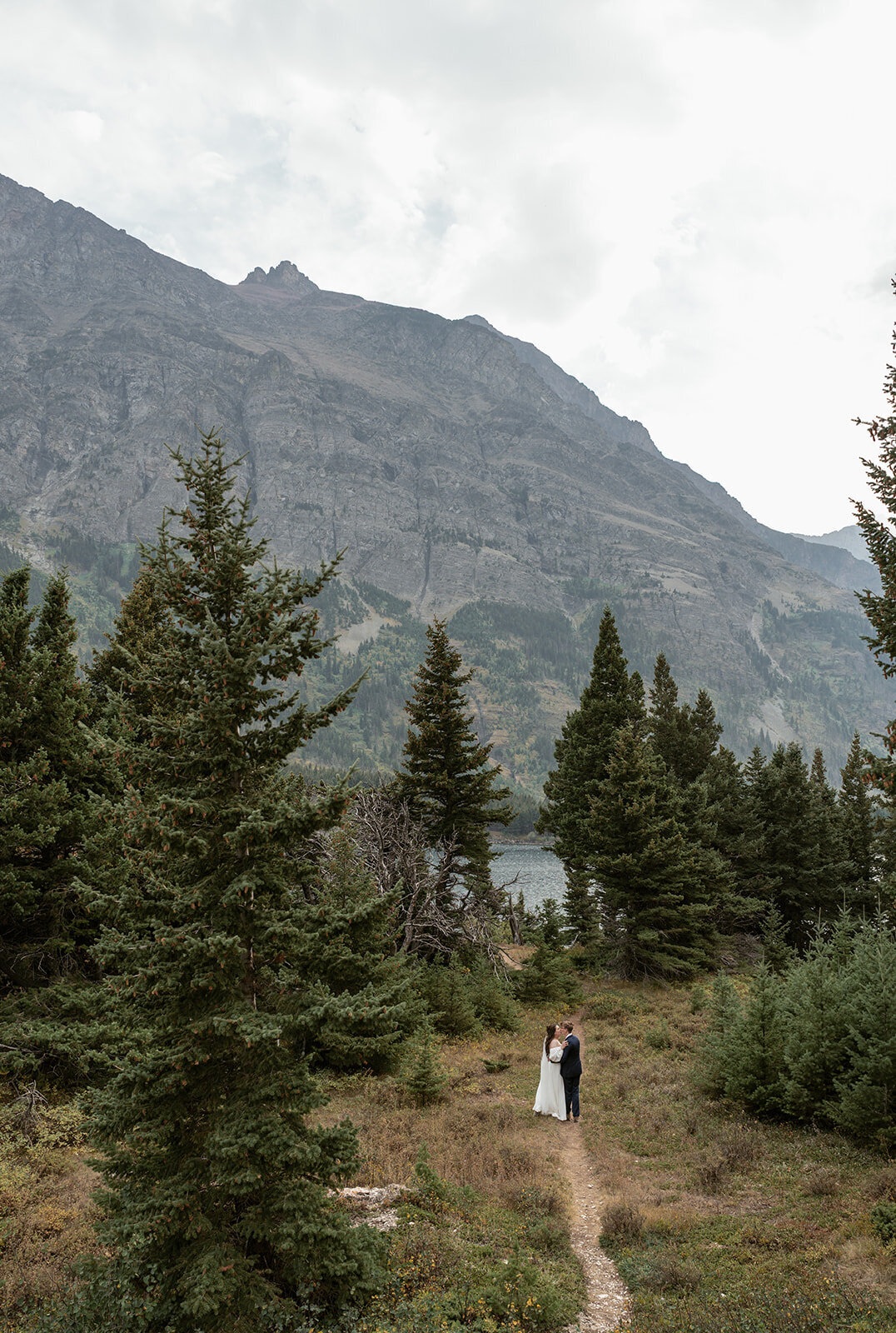 A couple shares an intimate moment surrounded by tall evergreens and towering mountain cliffs beside a lake in Glacier National Park, captured by Sydney Breann Photography during their Montana elopement.