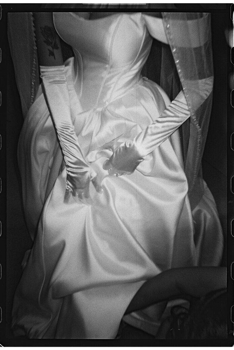 Black and white photo of a person wearing a satin dress, long gloves, and a veil, their hands clasped at their lap. A tattoo is visible on their shoulder. The composition is cropped tightly, focusing on the gown and hands.