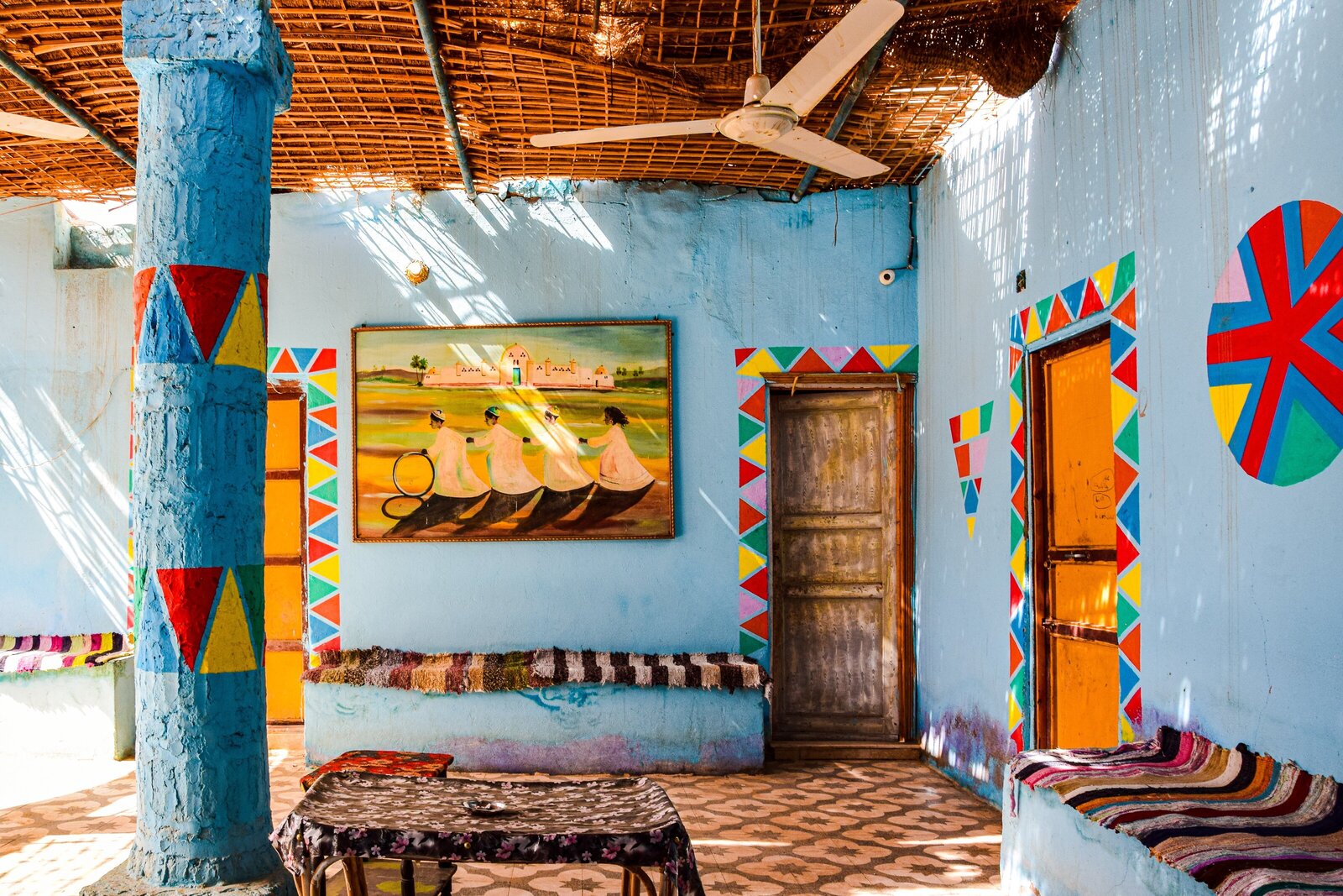 Interior of a Nubian village hom with thatch roof, light blue walls, wooden doors, and colorful painting on the wall and colorful red yellow and green banners painted around the door frames