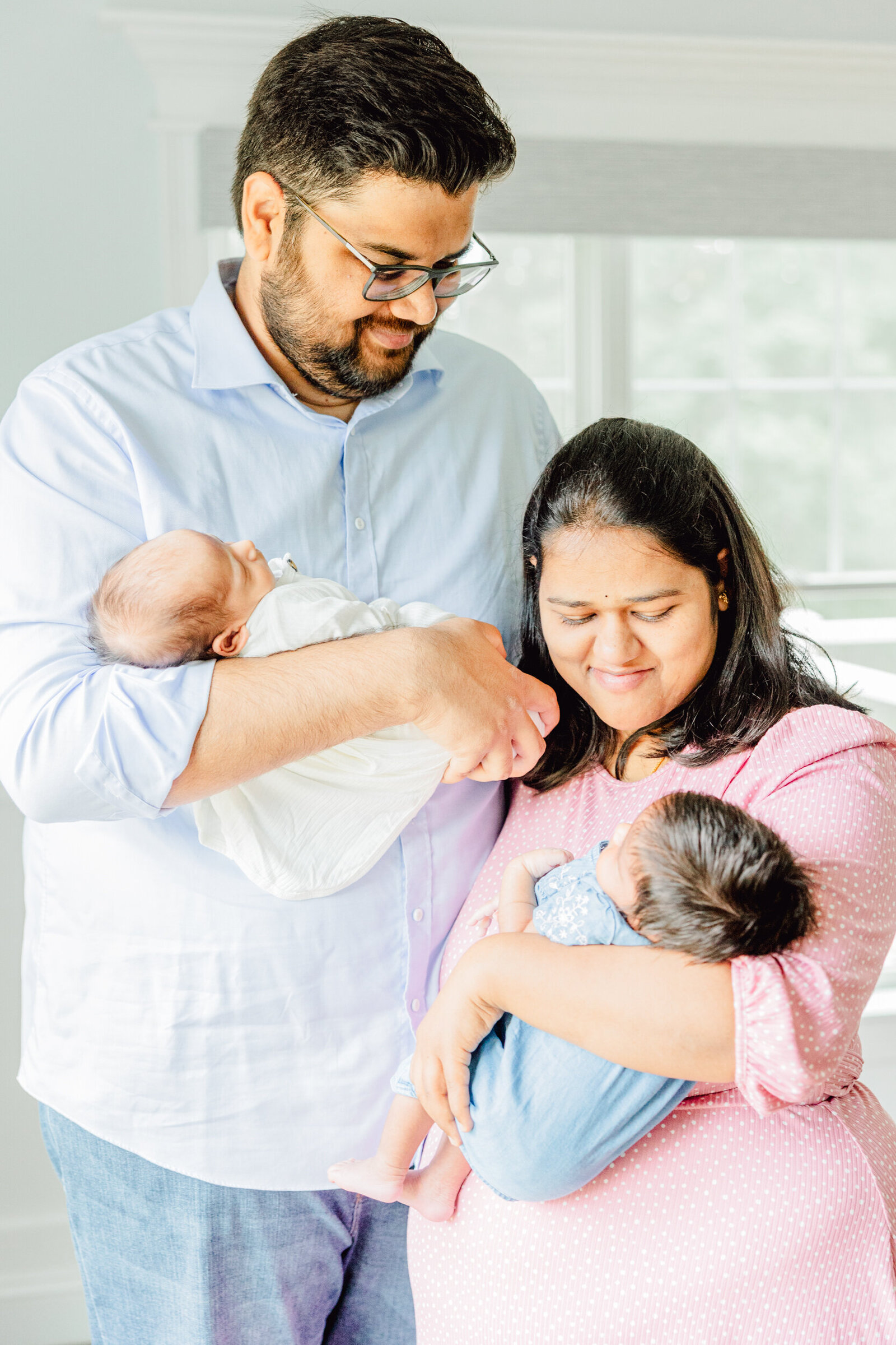 Mom and dad stand together in their living room, each holding a newborn and smiling at their babies