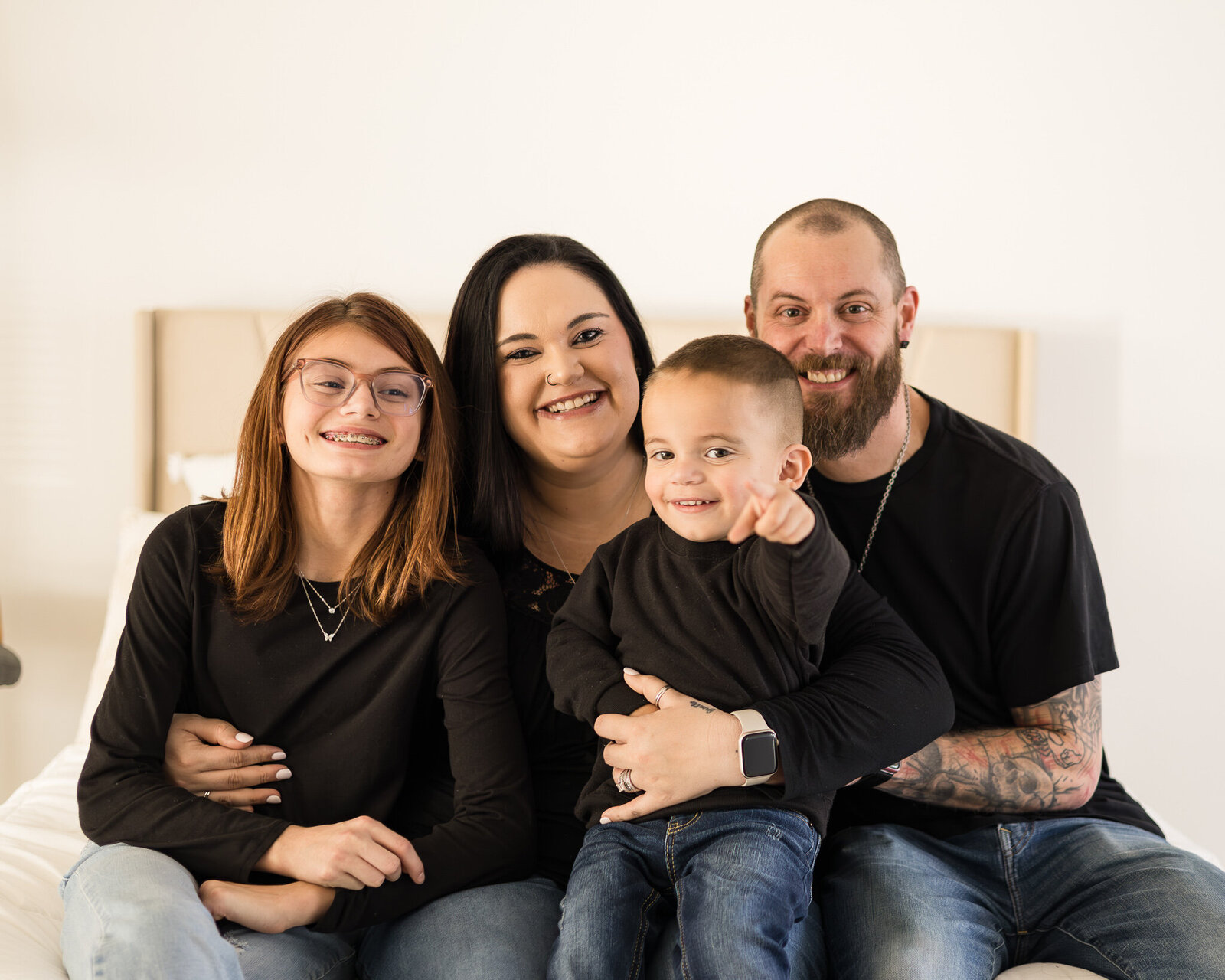 A family of 4 sitting on a bed looking at the camera smiling.  All of them are wearing black shirts and blue jeans.  Dad, son, and daughter have dark brown hair and mom has black hair