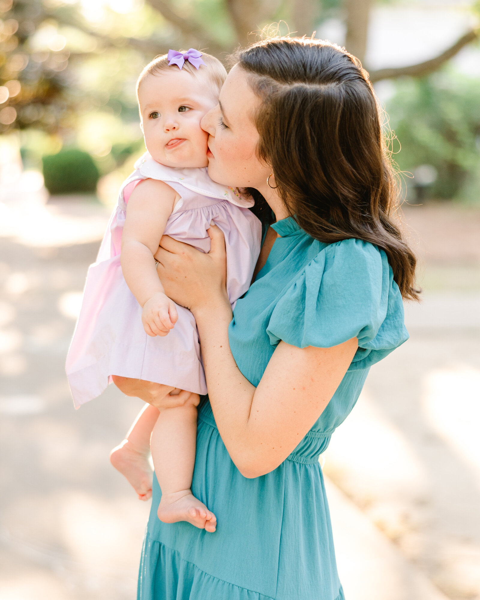 Mother kissing her baby in a lilac dress with a purple bow, surrounded by a sunny park, captured by Abby Bell Photography, Fine Art Portrait Photographer based in Alabama.