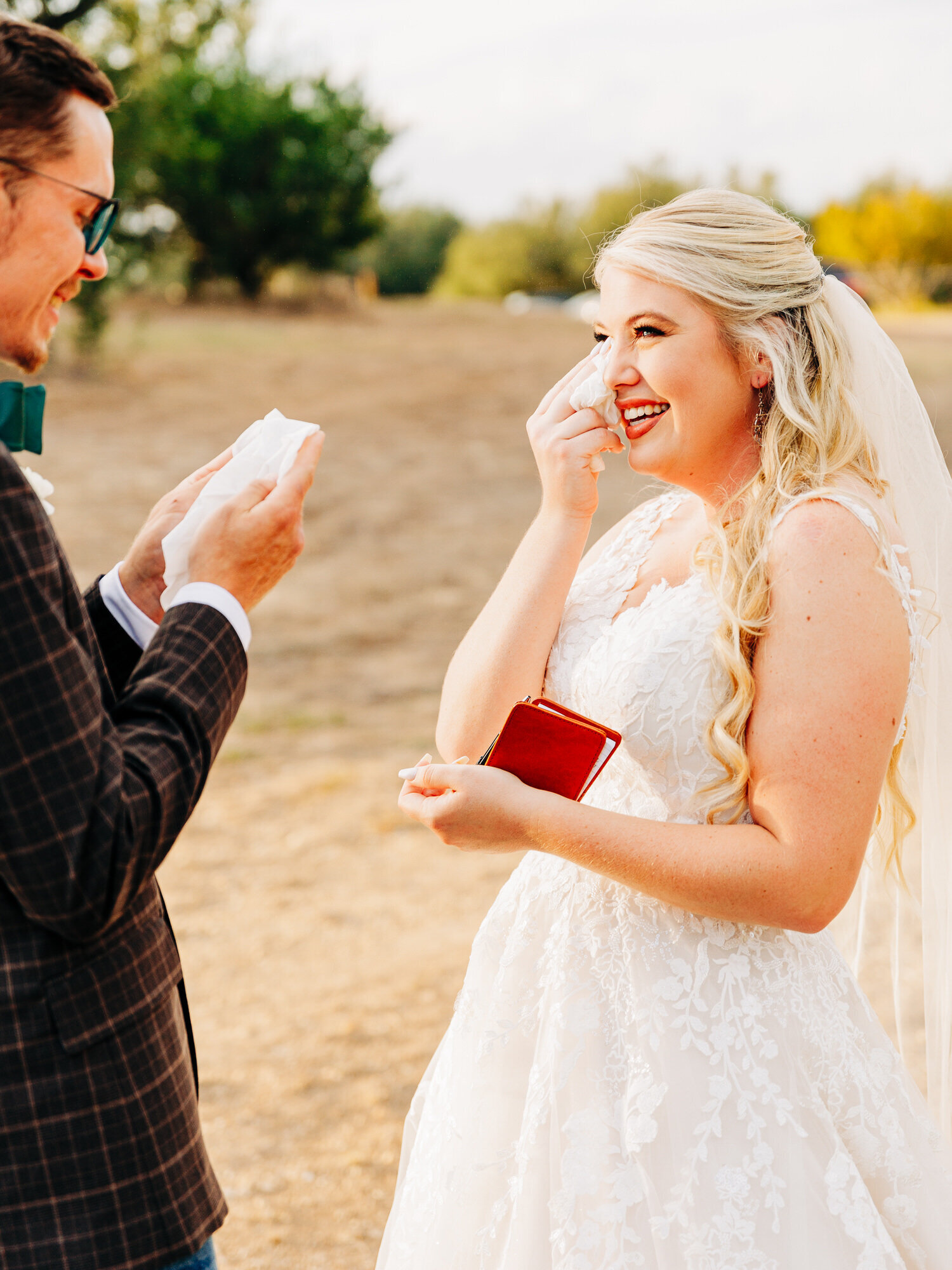 A bride in a white lace dress and veil wipes away happy tears while smiling at a man in a suit, who holds a tissue. Captured by an Eau Claire Wedding Photographer, they stand outdoors on a sunny day with trees and dry grass in the background.



